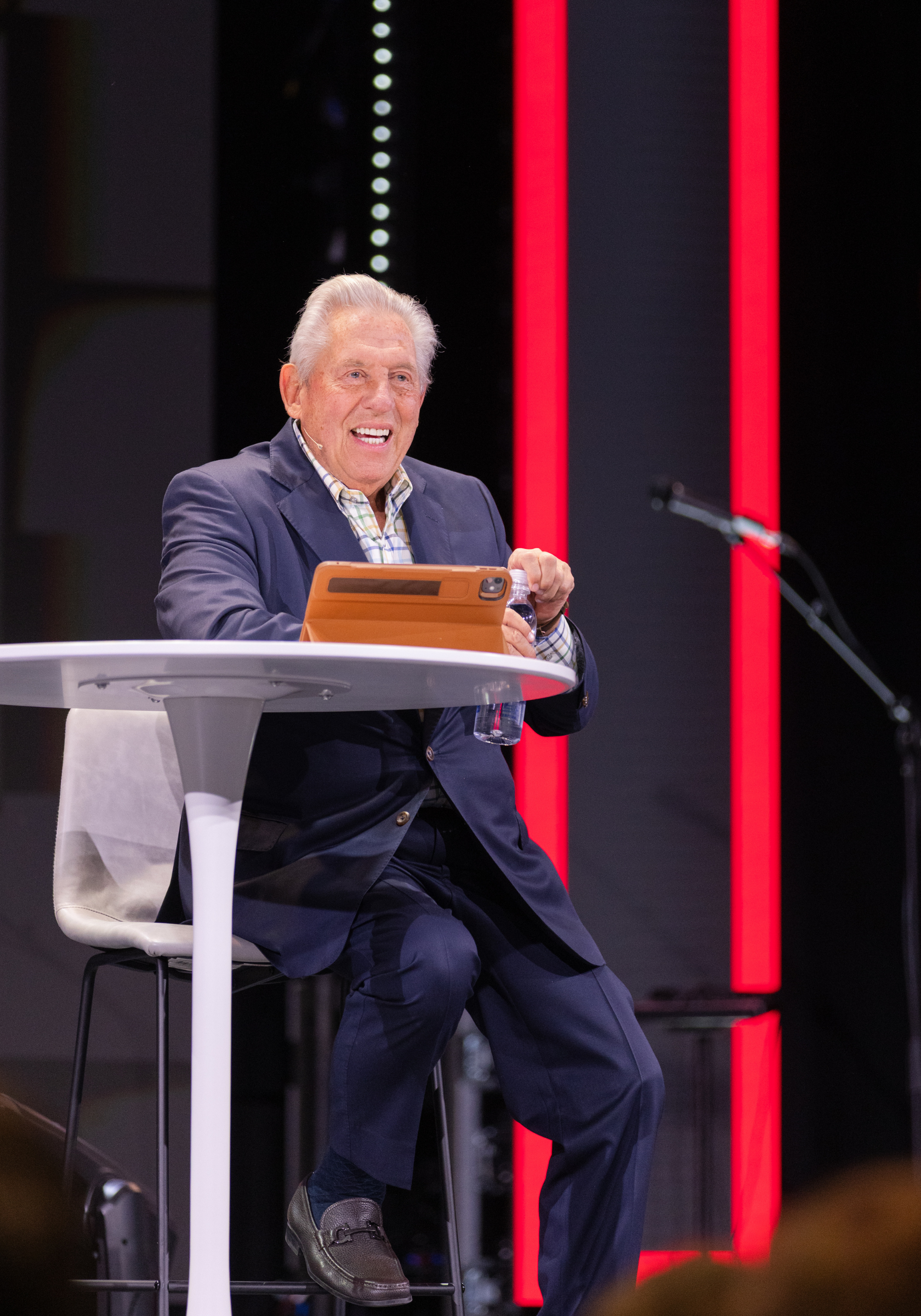 An elderly man with white hair, wearing a dark blue blazer and a checkered shirt, is sitting on a white chair at a round white table. He is smiling, holding a plastic water bottle in his right hand, and has a tablet with a tan cover mounted on the table in front of him. There is a microphone stand nearby, and the background features illuminated vertical red and black panels.