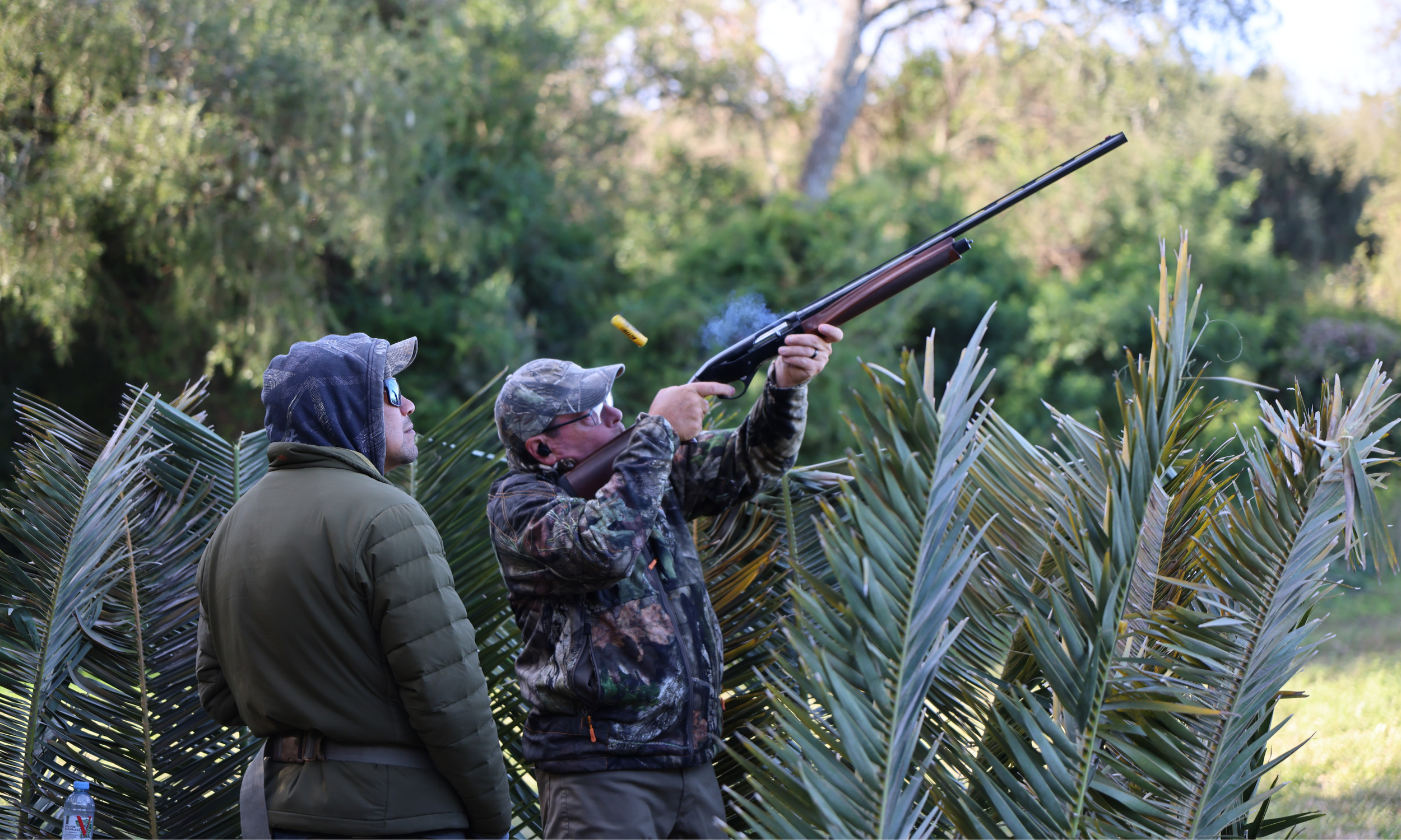 Two men, one wearing sunglasses, are outdoors, with one aiming and shooting a rifle in a forested area with tall, spiky plants.