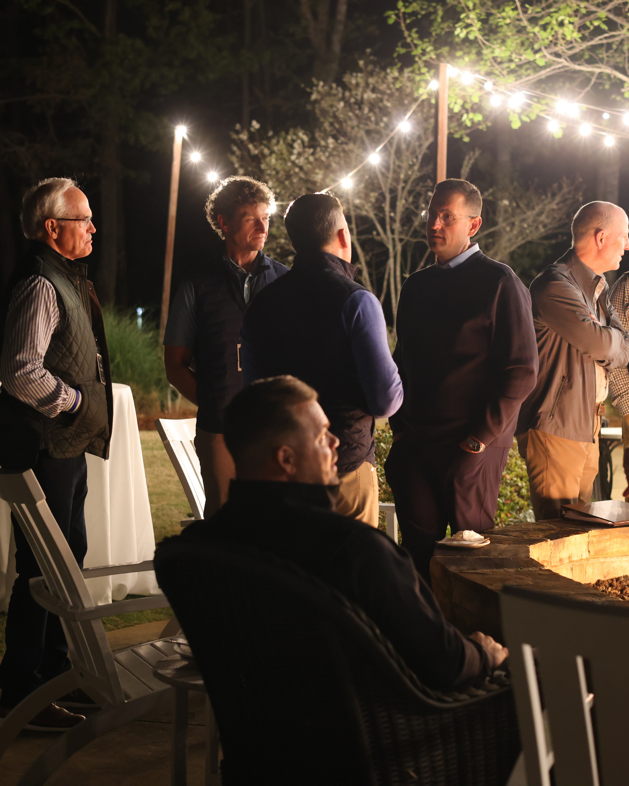 Group of men engaged in conversation at an outdoor gathering during nighttime, illuminated by string lights with trees in the background.