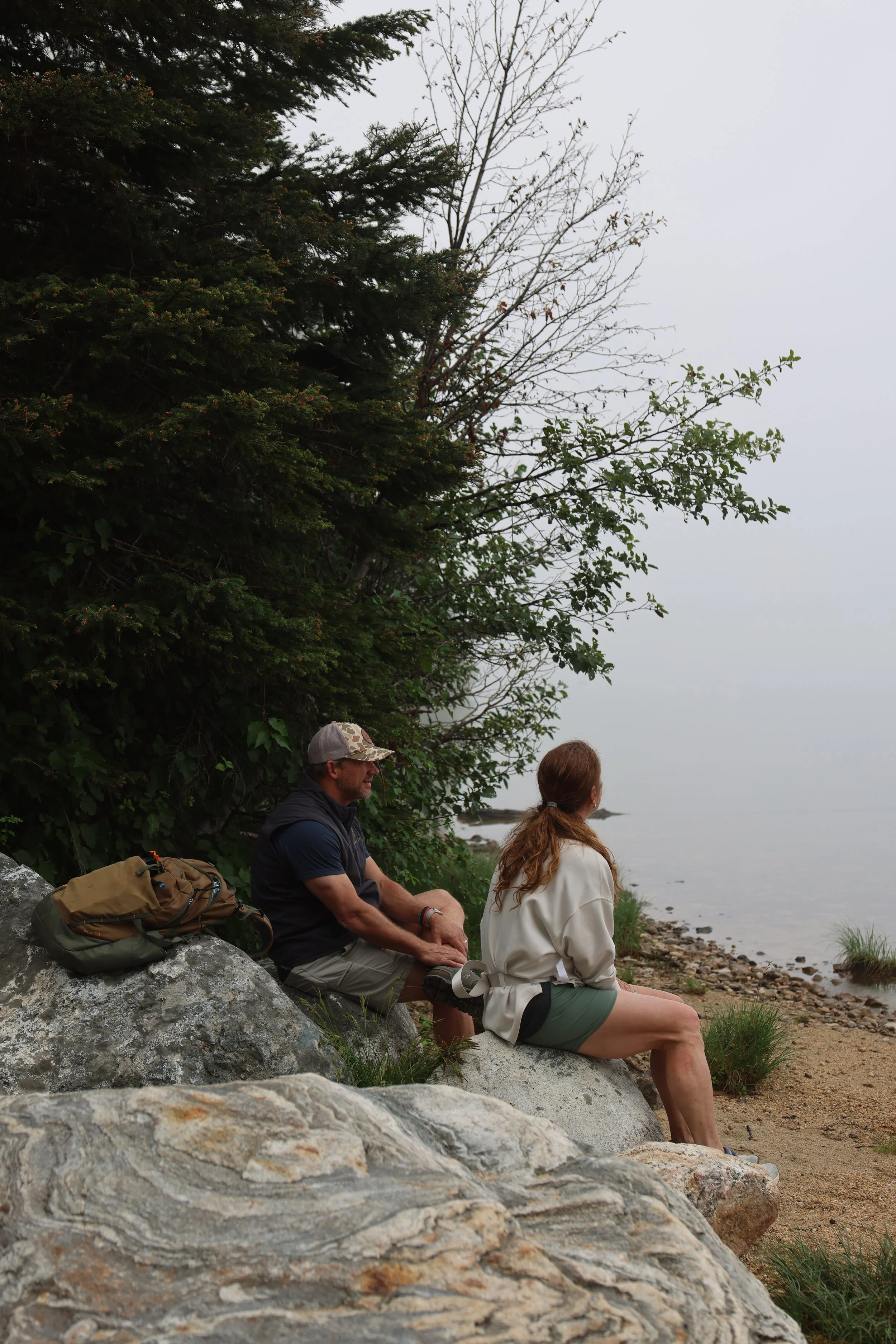 Two people sitting on rocks near the water's edge, with trees and rocky shoreline in the background.