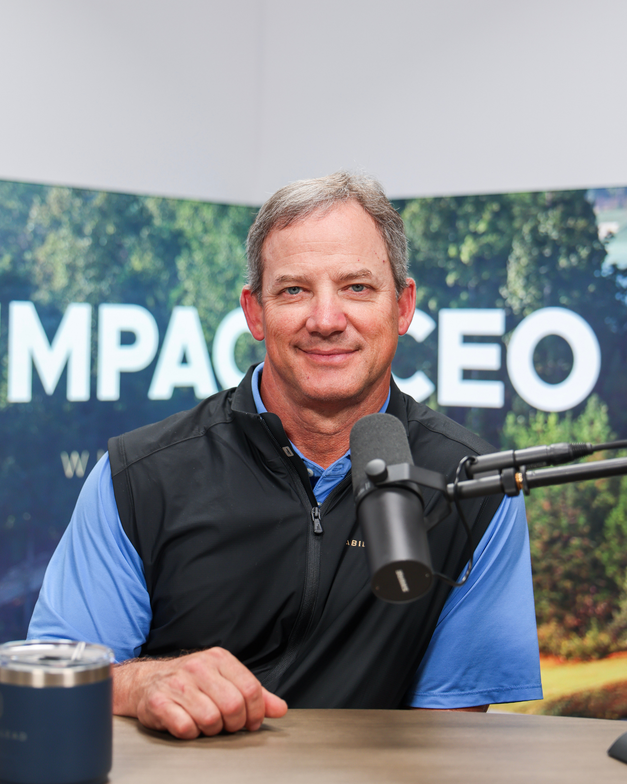 A man with gray hair and blue eyes sitting at a table with a black microphone in front of him, wearing a blue shirt and a black vest. His hand is resting on the table. In the background, part of a large sign with white letters and a green, wooded landscape is visible.