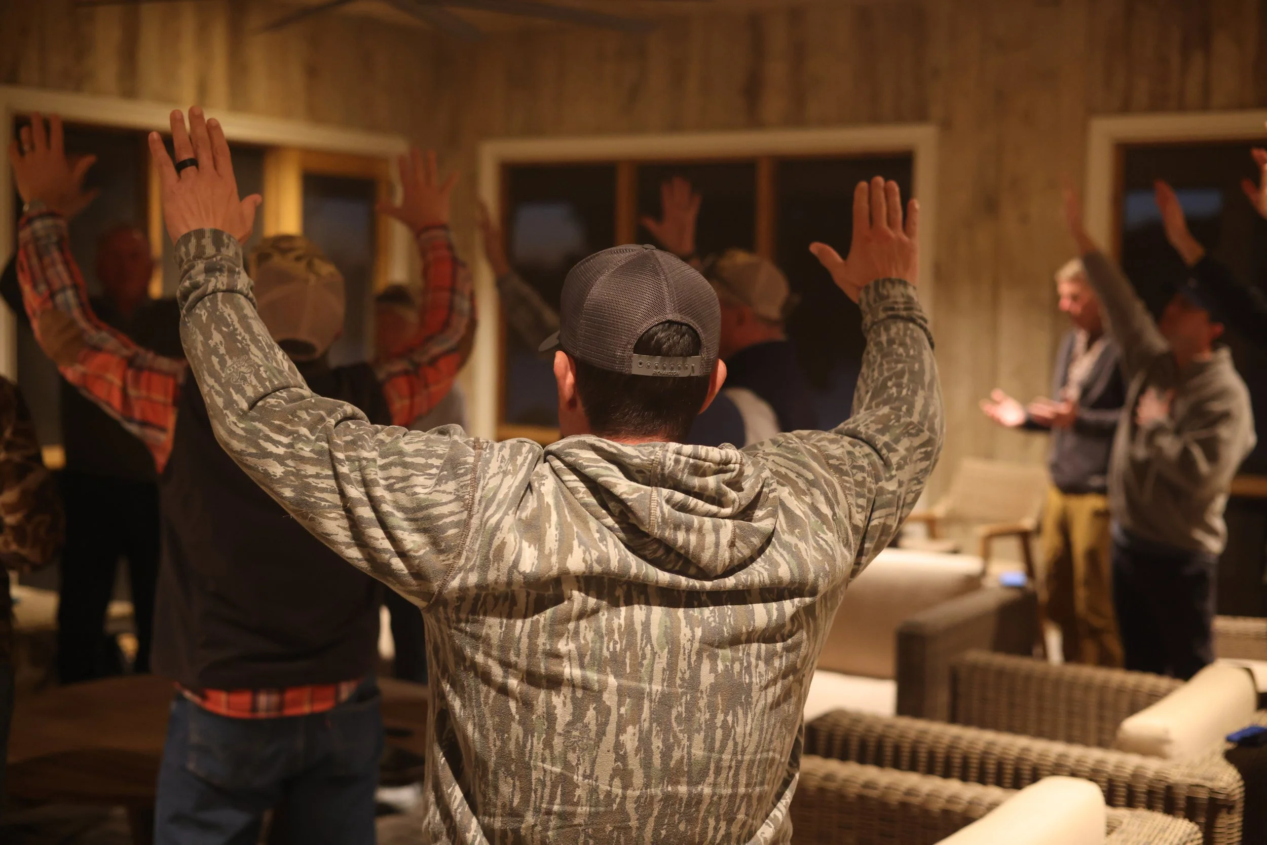 A group of people gathered indoors, raising their hands as part of a group activity or prayer. The focus is on a person in a camouflage hoodie and baseball cap with their back to the camera.