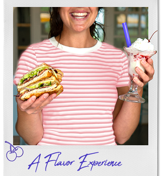 A person wearing a pink and white striped shirt holding one of our sandwiches in one hand and a milkshake topped with whipped cream and a cherry in the other, with a cherry graphic and the text "A Flavor Experience" at the bottom.