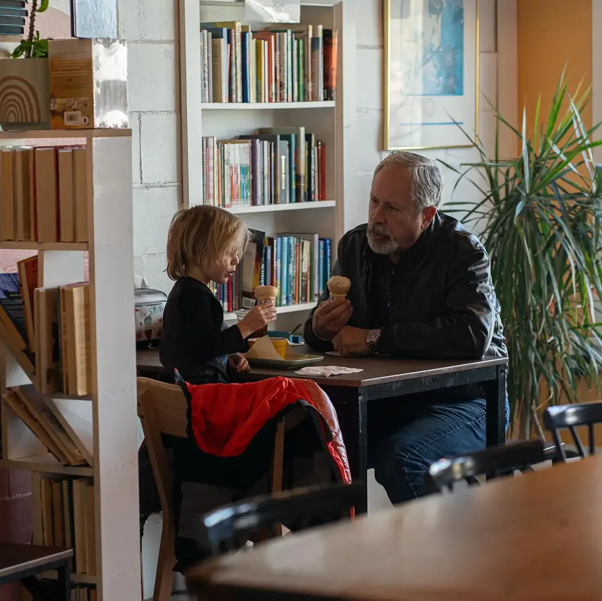 A man and a young boy sharing ice cream at a table in The Station at our Antrim location, a cozy, well-lit space with bookshelves and large windows.