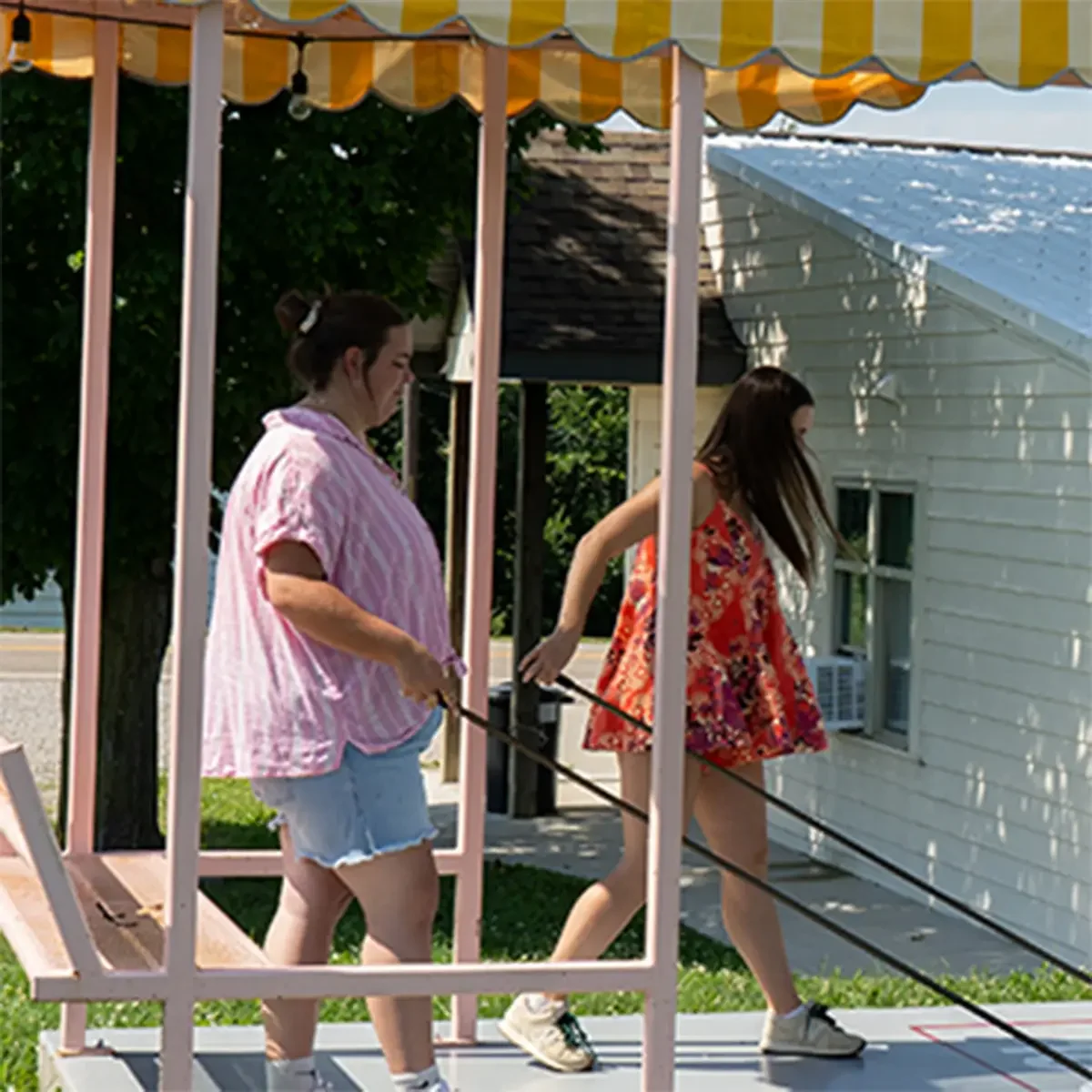 Two women walking on a mobile platform outdoors, one wearing a pink and white striped shirt and shorts, the other in a red floral dress.