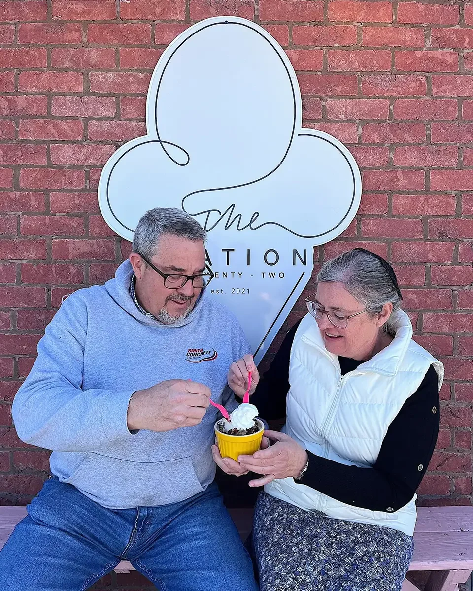 A man and a woman sitting on a bench, sharing a cup of ice cream with whipped cream, in front of a brick wall with a large sign reading 'The Celebration'.