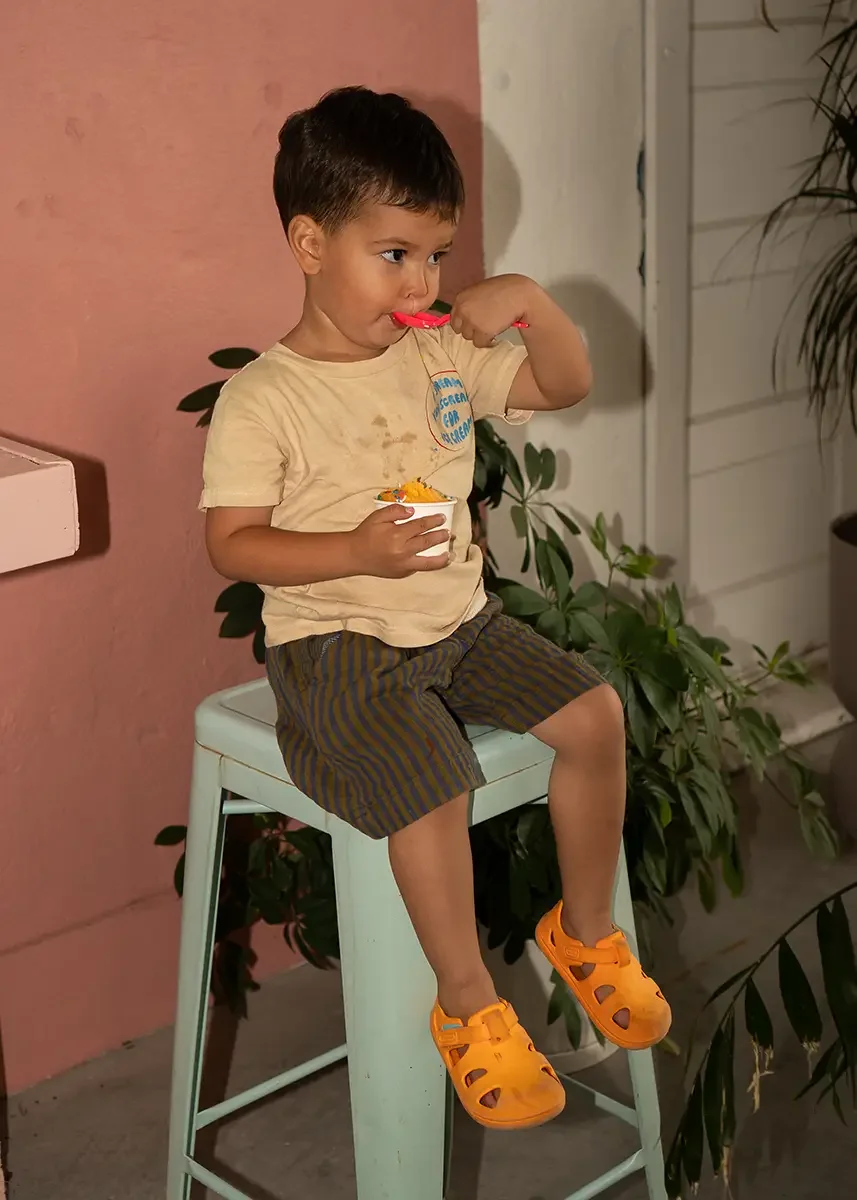 A young boy sitting on a high stool outdoors, wearing orange Crocs, eating ice cream with a plastic spoon, wearing a beige t-shirt and striped shorts, surrounded by some potted plants near a pink wall.