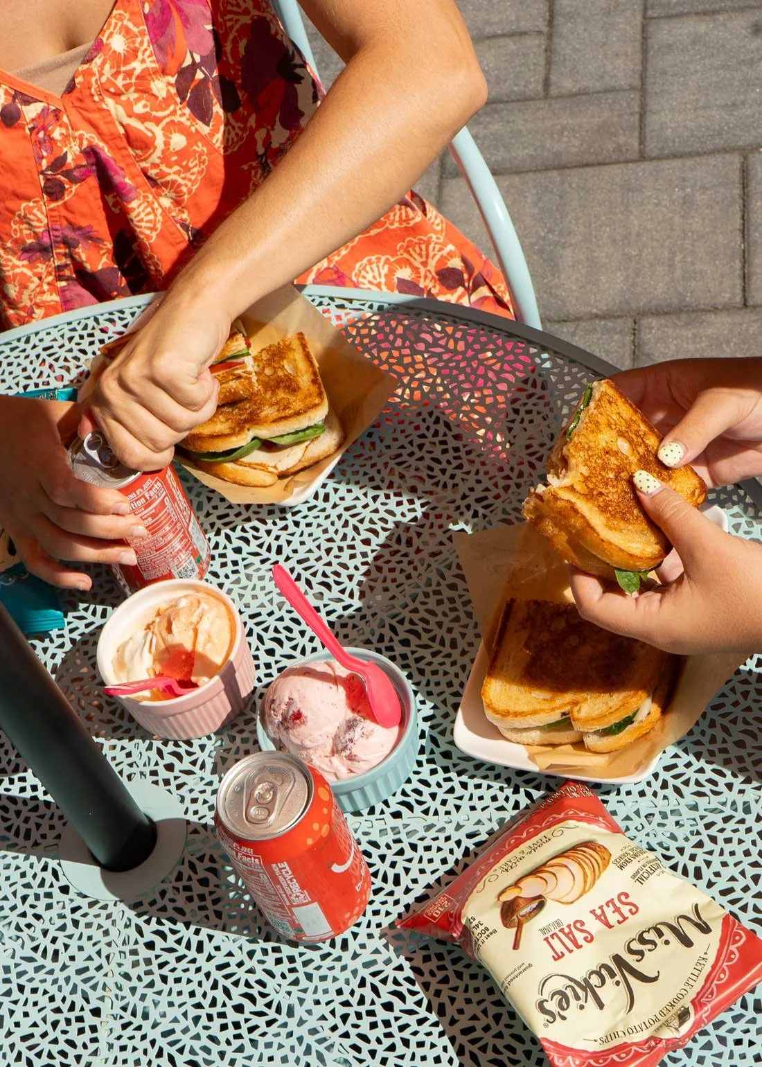 Two people eating sandwiches from The Station with pickles, sitting at a decorative table on our porch at our Antrim location. There are two cups of ice cream, a can of soda, a bag of snacks, and a bottle of soda on the table.