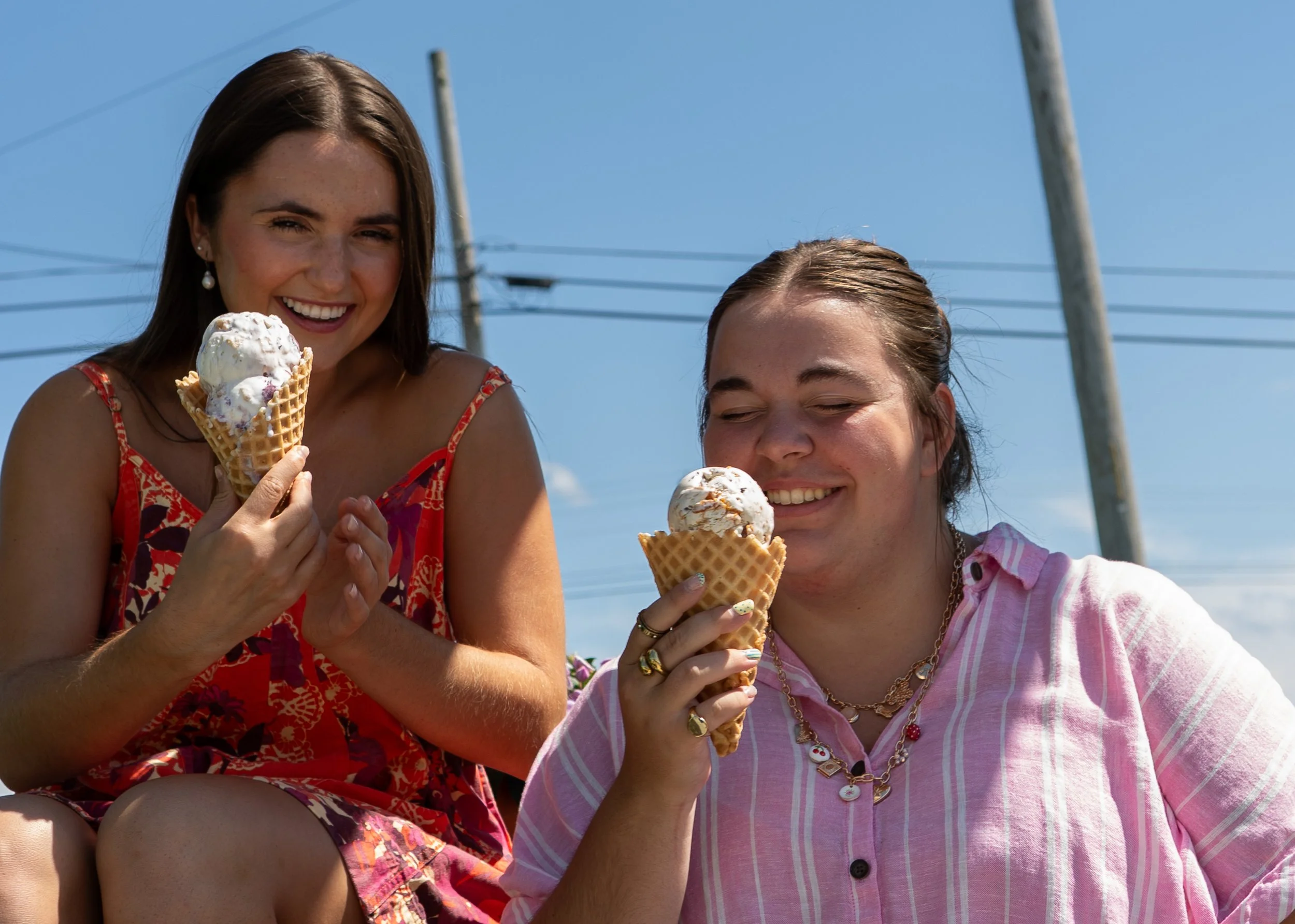 Two women enjoying ice cream cones outdoors on a sunny day, each with a Station best selling ice cream flavor: blueberry cheesecake on one waffle cone and triple peanut butter on the other.
