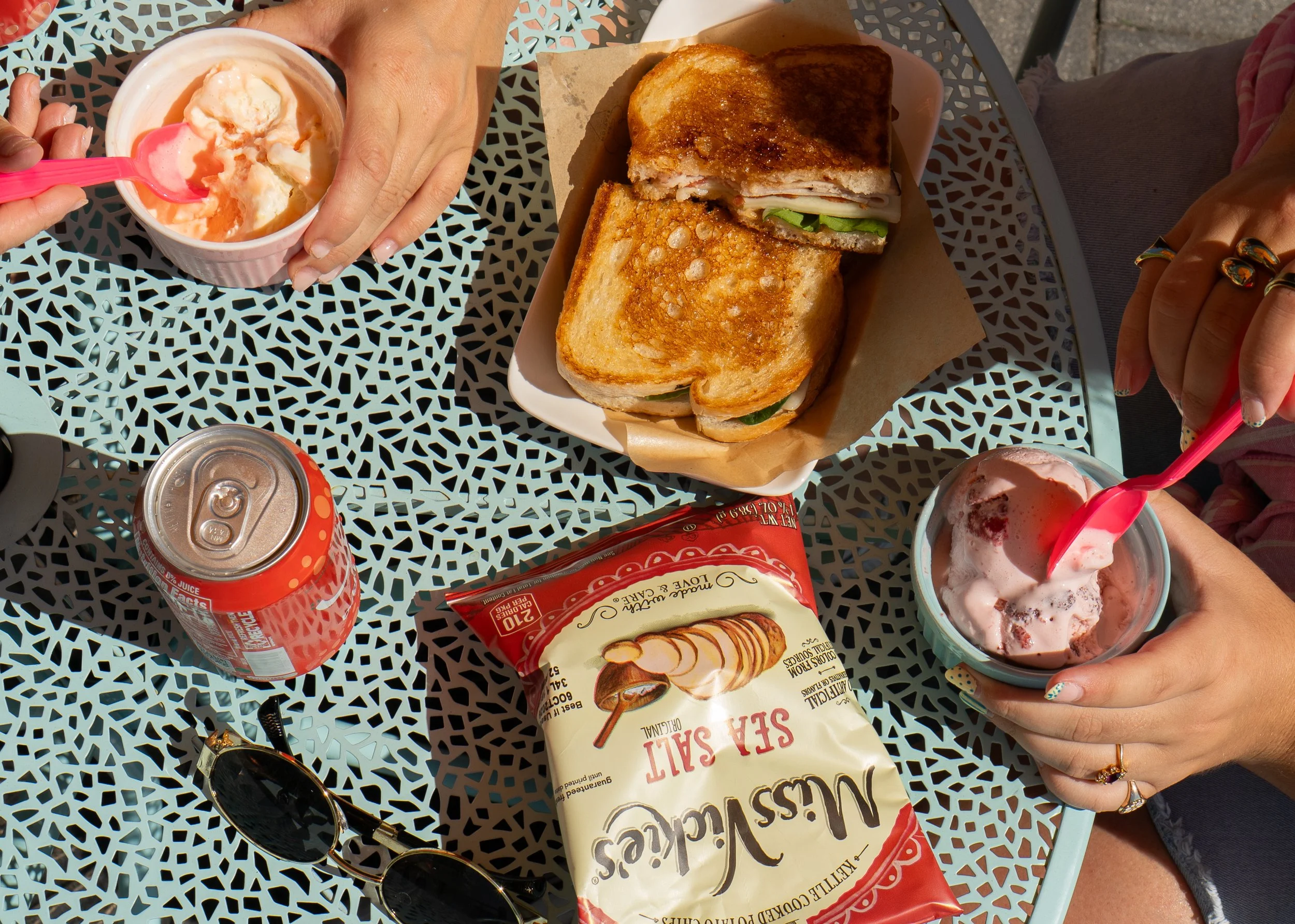 Two people enjoying lunch on The Station patio. A sandwich with toasted bread, lettuce, and cold cuts, a cup of vanilla ice cream with a pink spoon, a can of soda, and a bag of sea salt potato chips are seen. 