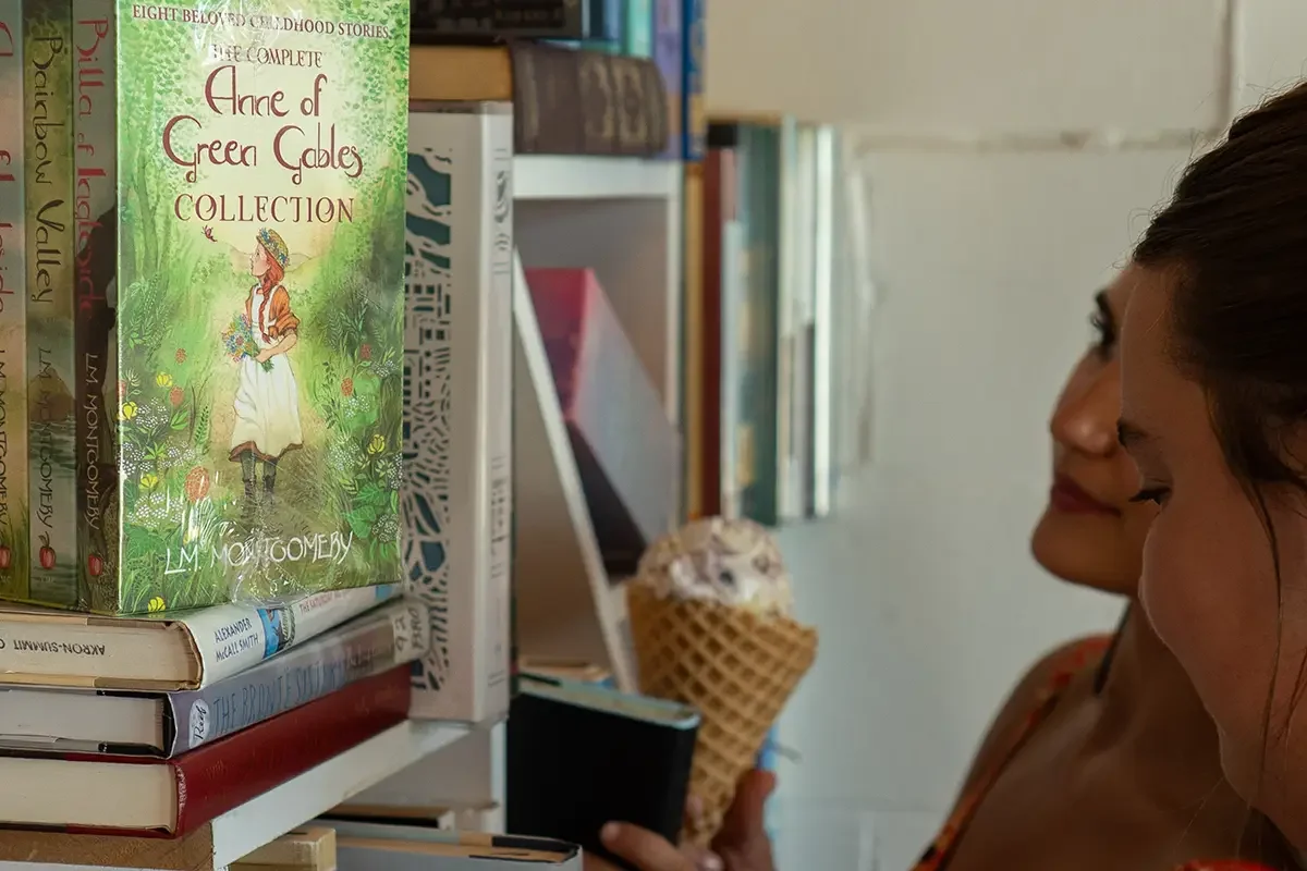 Two women are browsing our books at The Station. One woman is holding an ice cream cone, and the other woman is looking at a book titled 'Anne of Green Gables Collection' by L.M. Montgomery.