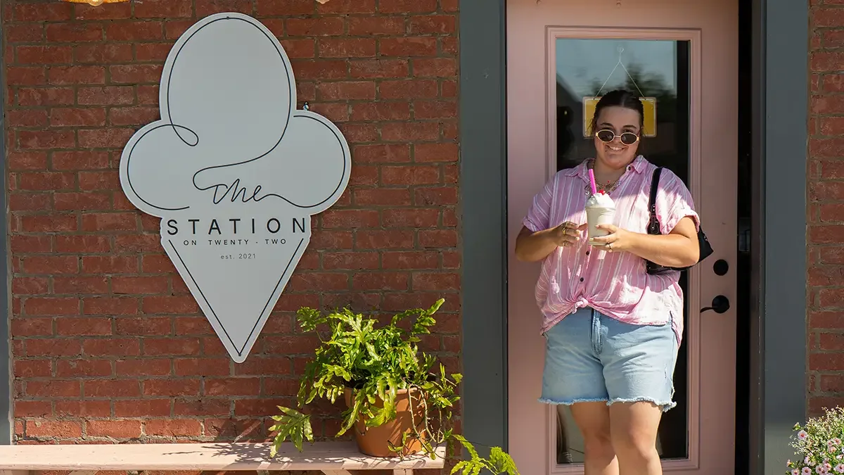 A woman with sunglasses, wearing a pink striped shirt and denim shorts, is standing outside a pink door at The Station, holding two milkshakes. To her left, there is The Station sign with our logo, and a potted plant is below the sign.