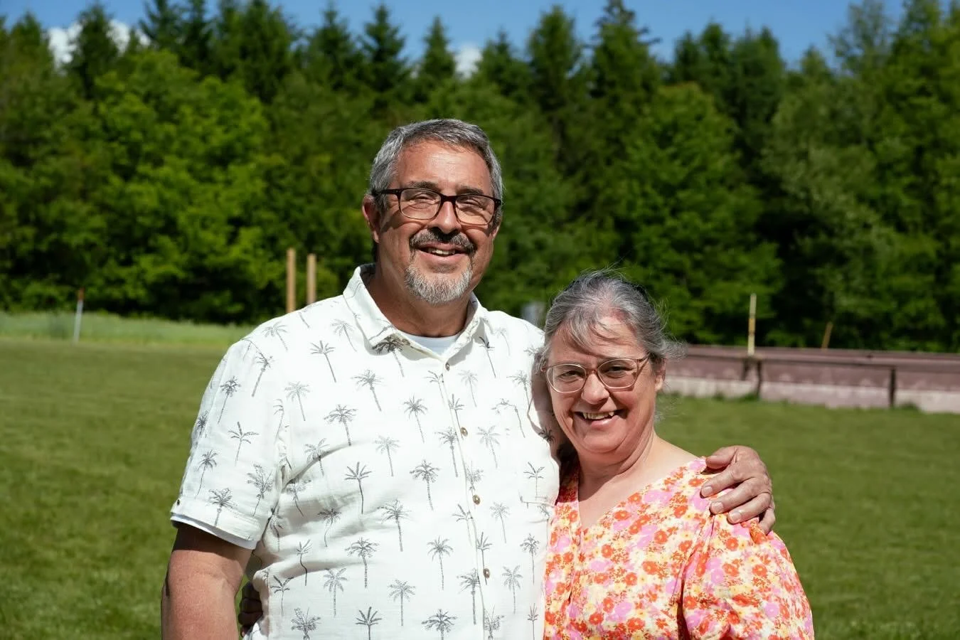 This smiling couple, The Station owner, Ron Miller and his wife, Ann, are standing outdoors on a sunny day with a background of green trees and grass.