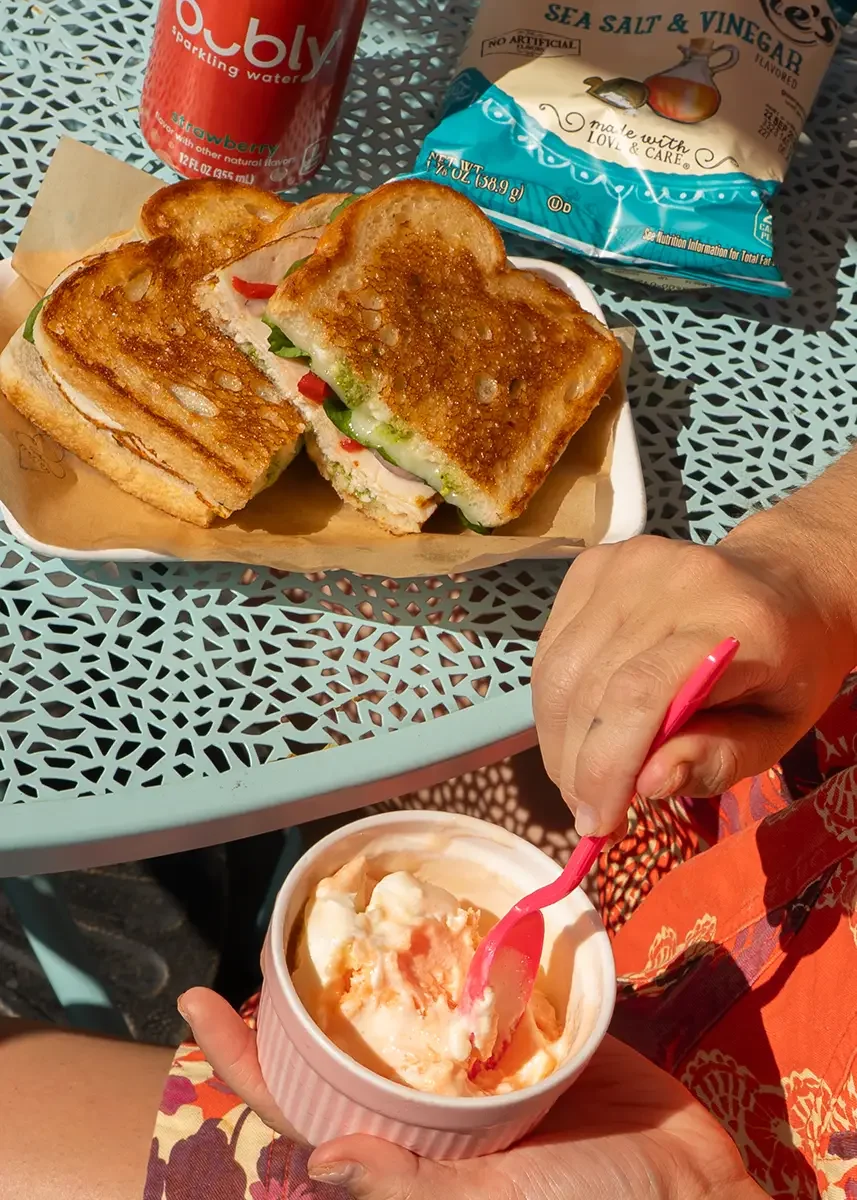 A plate with two grilled sandwiches containing turkey, lettuce, and tomato, a can of strawberry sparkling water, a bag of sea salt and vinegar potato chips, and a person holding a container of vanilla ice cream with caramel syrup. The setting is an o