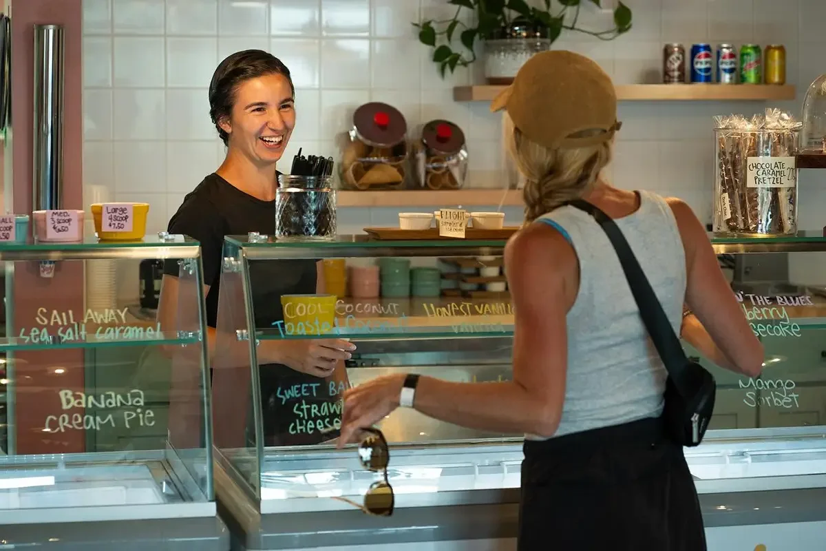 A woman with long hair, wearing a tank top with a strap, and carrying a black shoulder bag, is ordering ice cream from one of our smiling staff members at The Station. The shop has various colorful signs and flavors listed on the glass display.