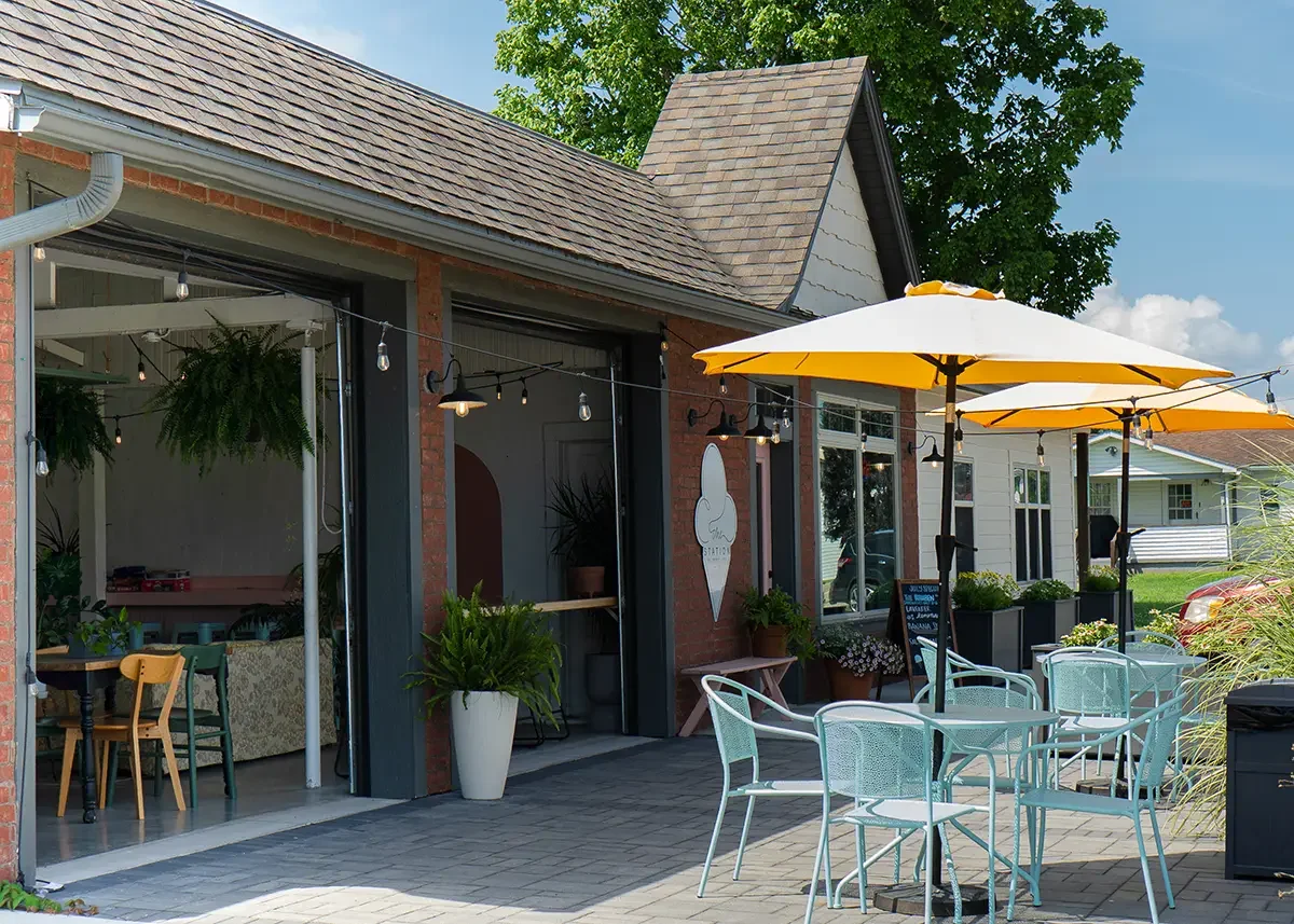 Our outdoor patio with yellow umbrellas, turquoise metal tables and chairs, potted plants, and The Station brick building set in the background with string lights hanging above.