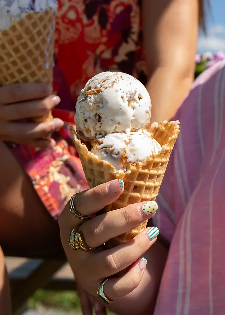 Close-up of a hand holding a waffle cone with two scoops of vanilla ice cream topped with caramel sauce, with people in summer clothing and a bright outdoor setting visible in the background.