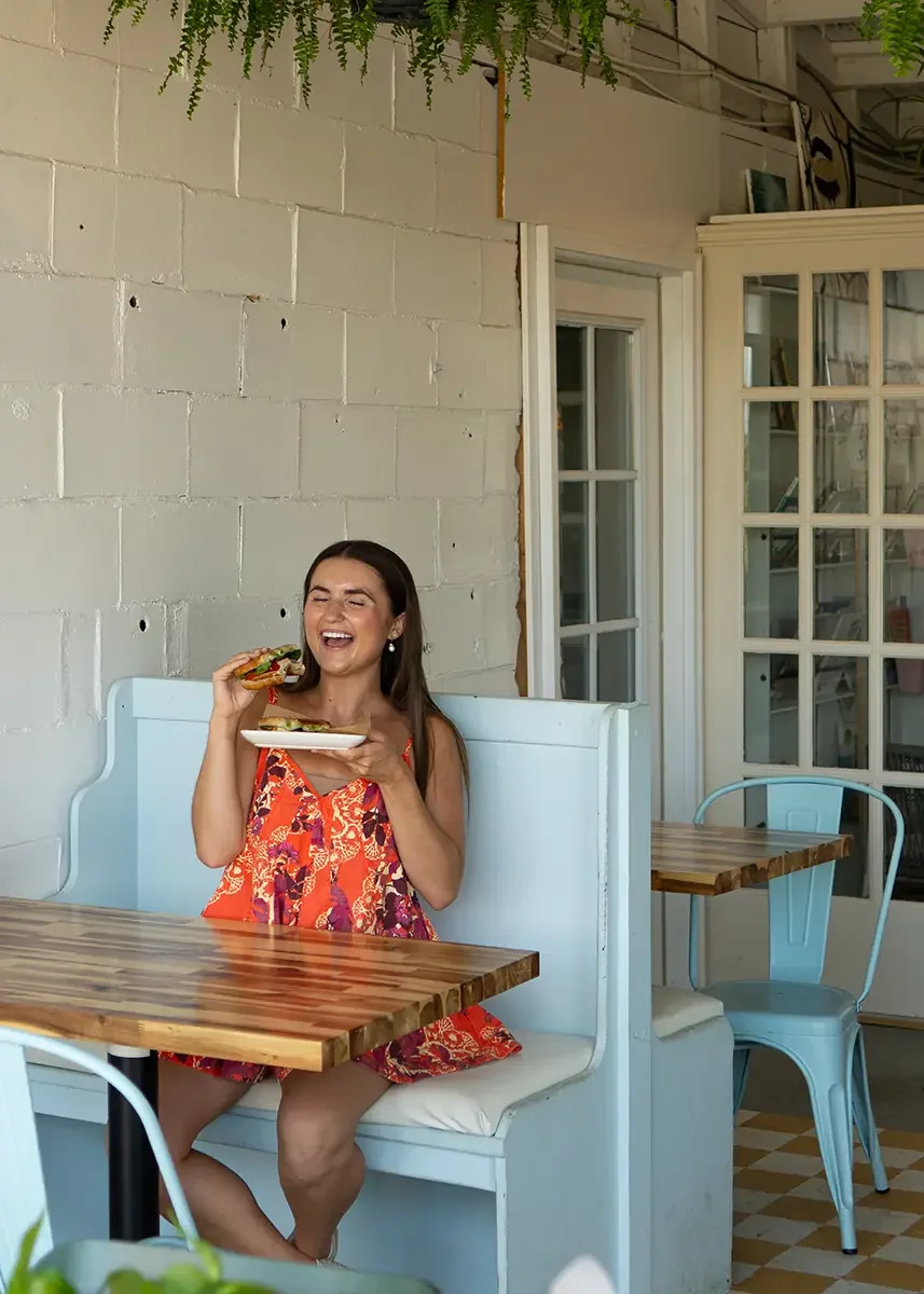 A woman in a colorful dress is seated at a Station booth, holding a plate with one of our sandwiches, laughing, and enjoying her meal.