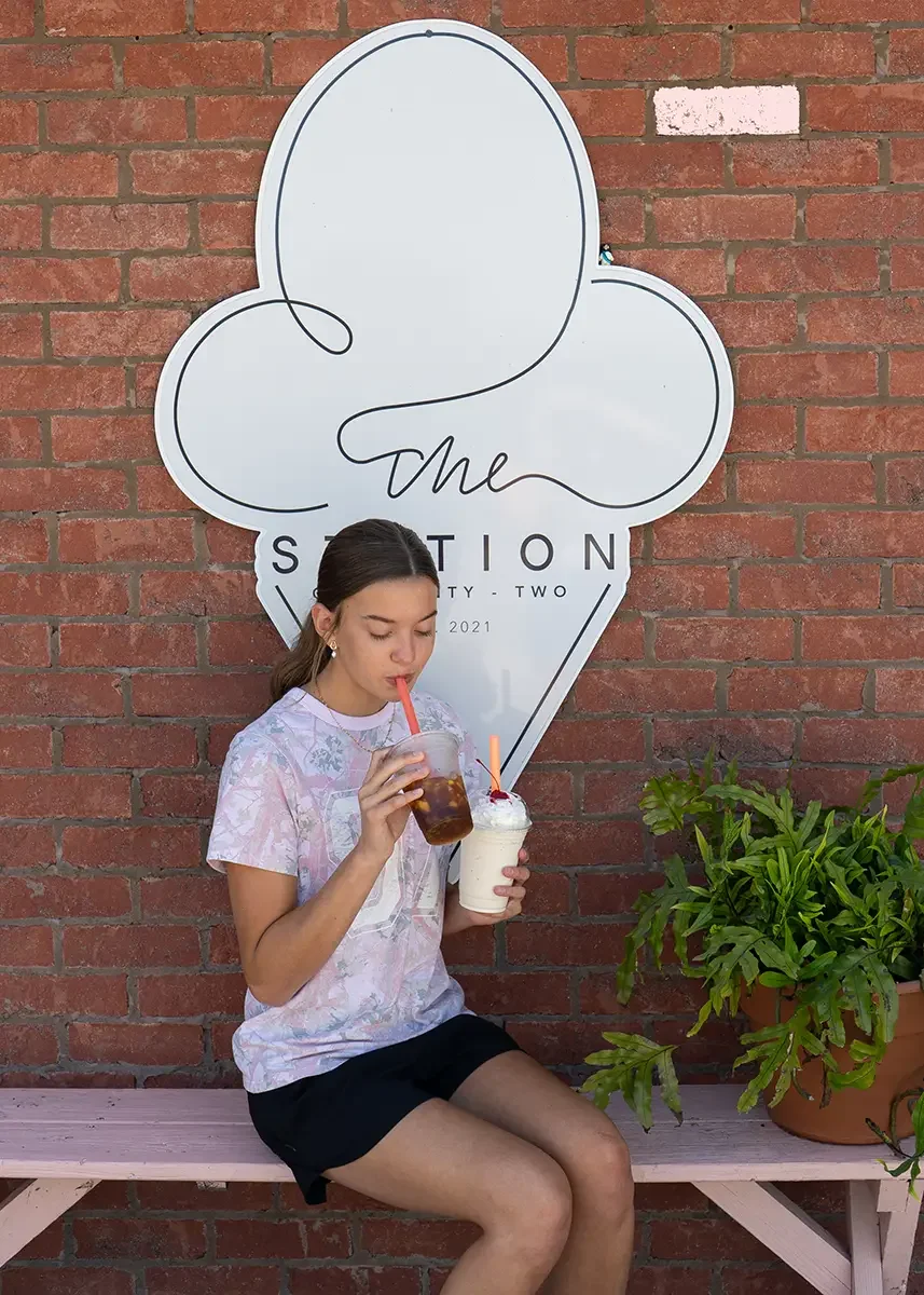 A young woman sitting on a pink wooden bench, sipping an iced coffee and a milkshake, in front of The Station sign and a potted plant nearby.