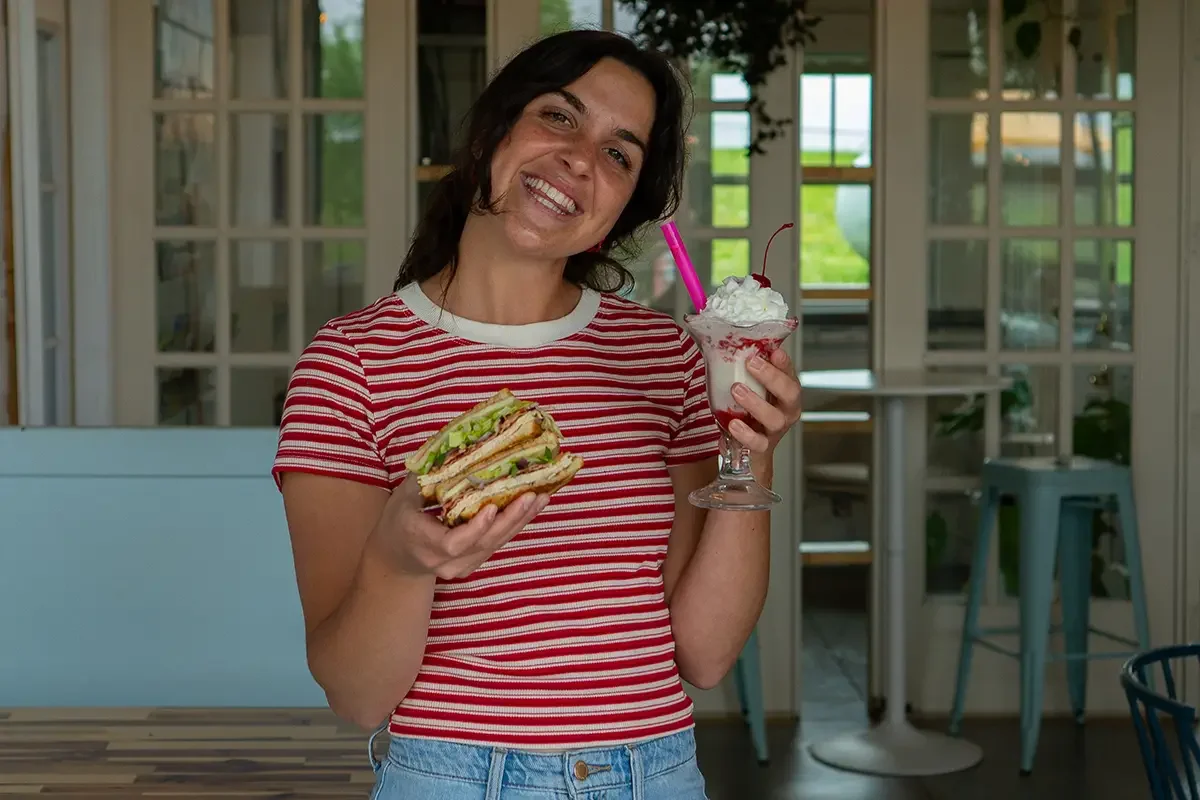 A woman smiling while holding one of The Station sandwiches in one hand and an ice cream sundae in the other, inside The Station garage space with glass-paneled doors and green trees outside.