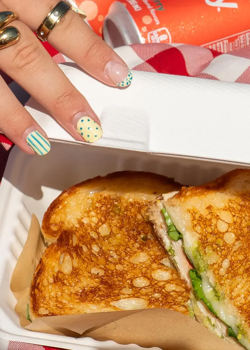 Close-up on one of The Station's monthly special sandwiches: toasted bread, lettuce, and turkey, inside a white takeout container. In the background, there is a red and white checkered napkin and a can of orange soda.