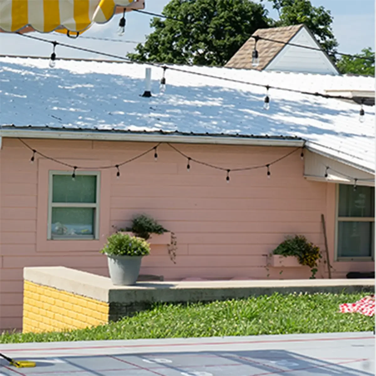 Exterior of a pink house with string lights hanging above. The house has two windows, and a planter box with yellow flowers on the right side. A concrete ledge with a potted plant is in the foreground, with a grassy area and a red-and-white checkered cloth on the right.