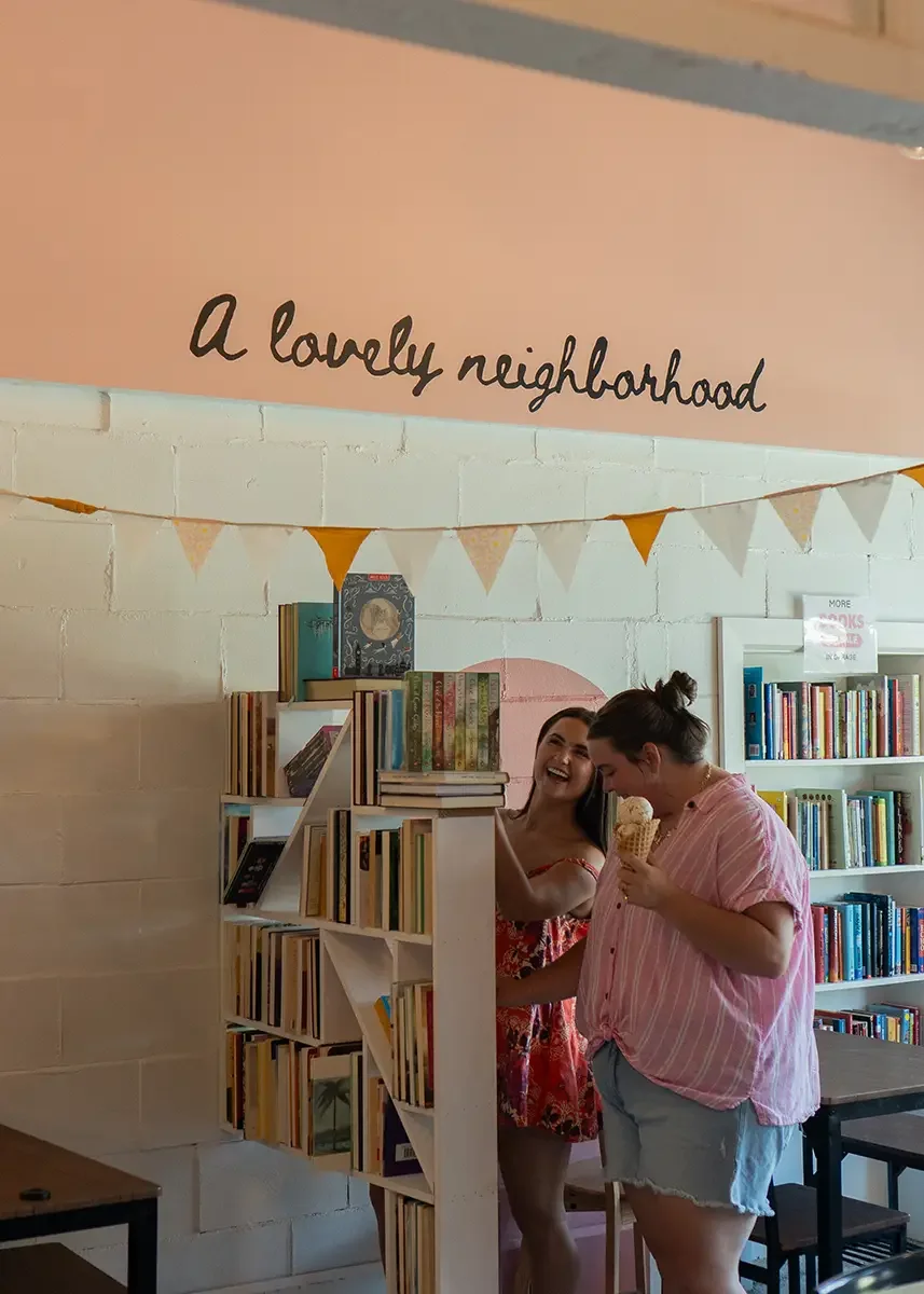 Two women enjoying ice cream inside a bookstore or library with a shelf of books, a pink wall with the phrase 'A lovely neighborhood' written on it, and decorative bunting hanging above.