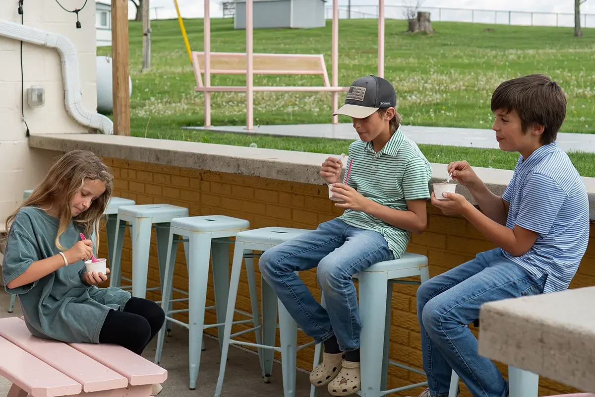 Three children sitting outside on stools, eating ice cream bowls. One girl in a green dress and black leggings, and two boys in striped shirts and jeans.