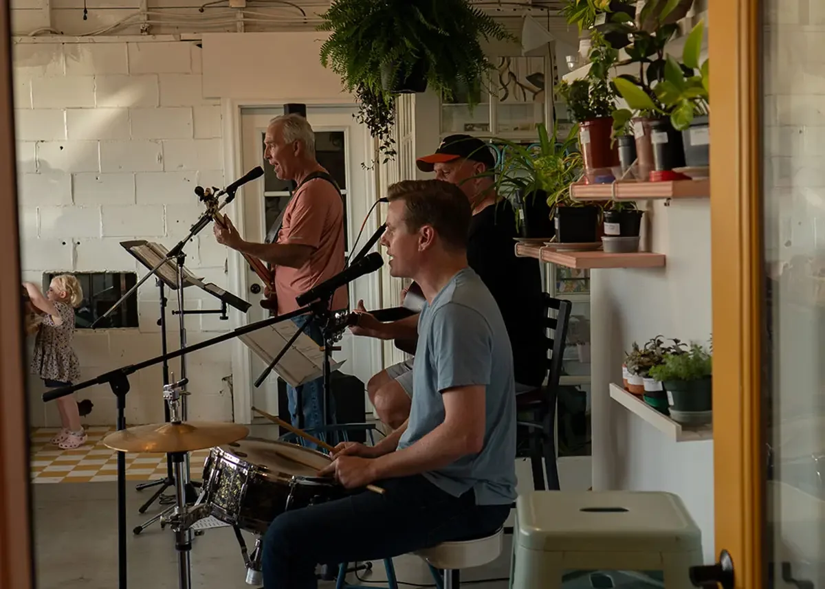 A band of three musicians performing indoors with plants on shelves and a white brick wall in the background, while a little girl in a dress and crocs observes from the side.