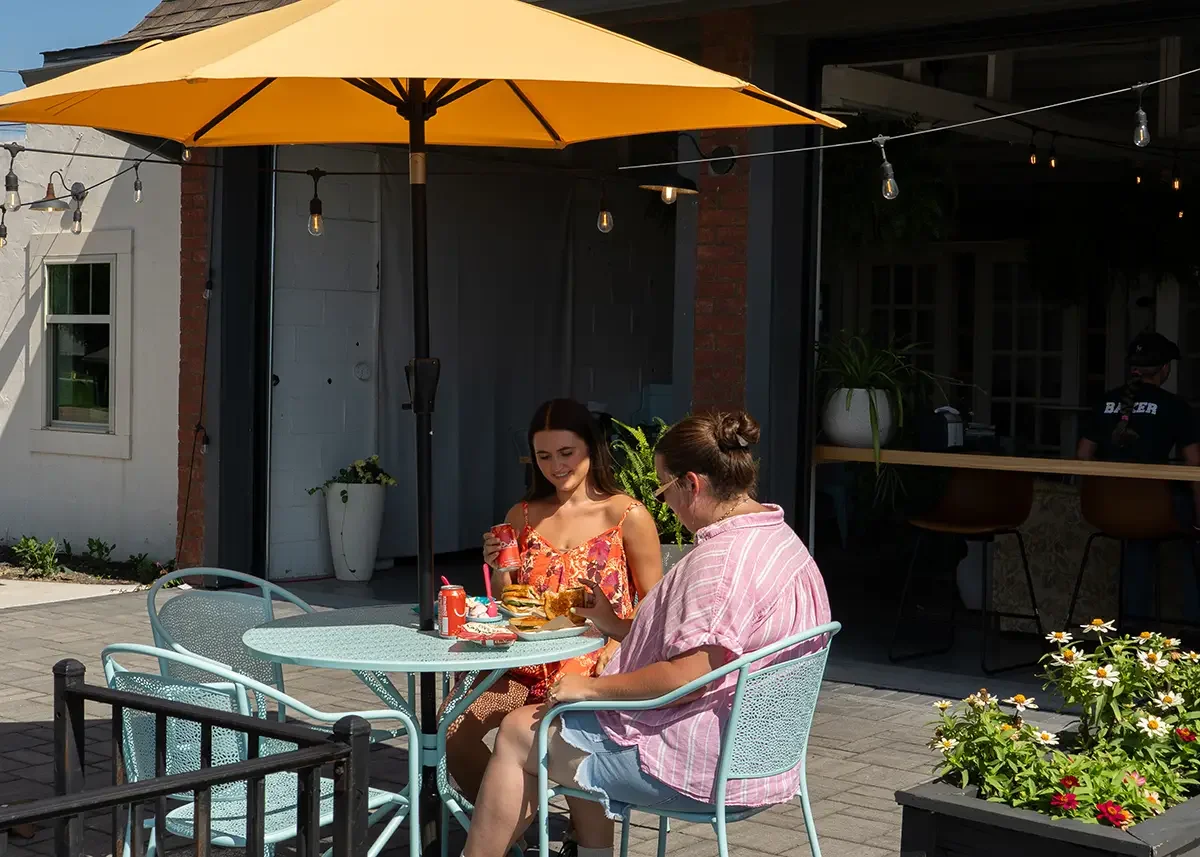 Two friends sitting at a patio table under a large yellow umbrella at The Station, our Antrim location, enjoying food and drinks, with a building and hanging string lights in the background.