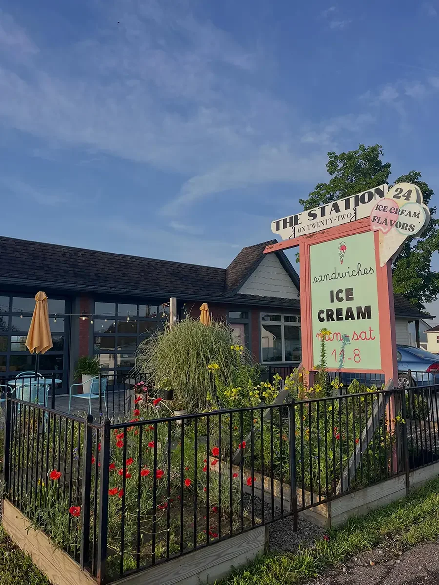 A small garden with colorful flowers sits in from of The Station exterior building at our Antrim location with large windows and a patio with umbrellas. A sign on a post advertises our ice cream and sandwiches, with a blue sky overhead.