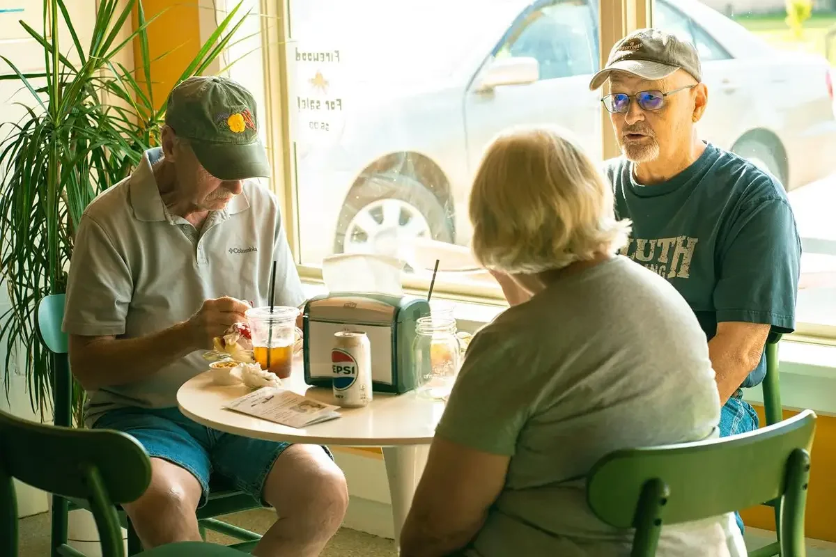 Three seniors sitting at a table inside The Station by our window, engaged in conversation, one man is having our lunch special, and a large plant is visible in the background.