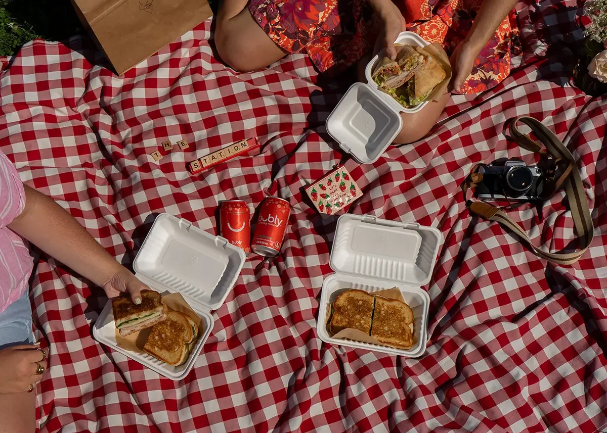 Picnic on a red and white checkered blanket with sandwiches in takeout boxes, cans of soda, a camera, and Scrabble tiles spelling 'THE STATION'.