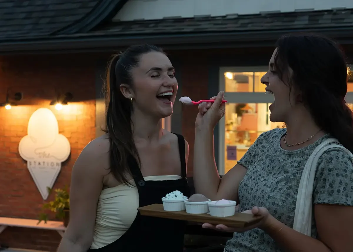 Two women share The Station ice cream samples outdoors in the evening, with one feeding the other from a spoon, holding a tray of different ice cream flavors, in front of The Station.