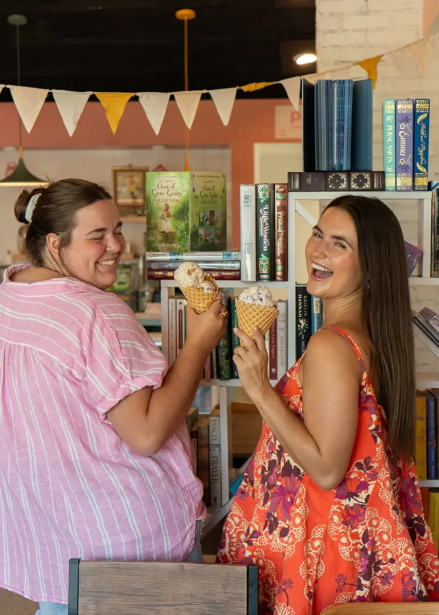 Two women with ice cream cones enjoying dessert at a bookstore or library, with shelves of books behind them and festive bunting hanging above.