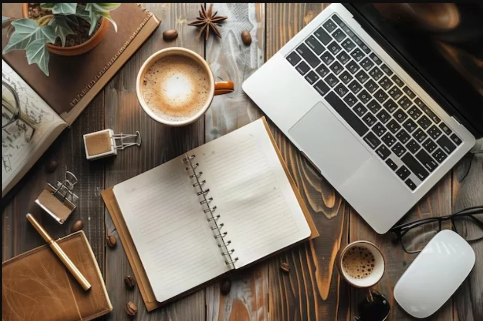A workspace on a wooden desk with a laptop, open notebook, cups of coffee, glasses, a plant, a book, and various office supplies.
