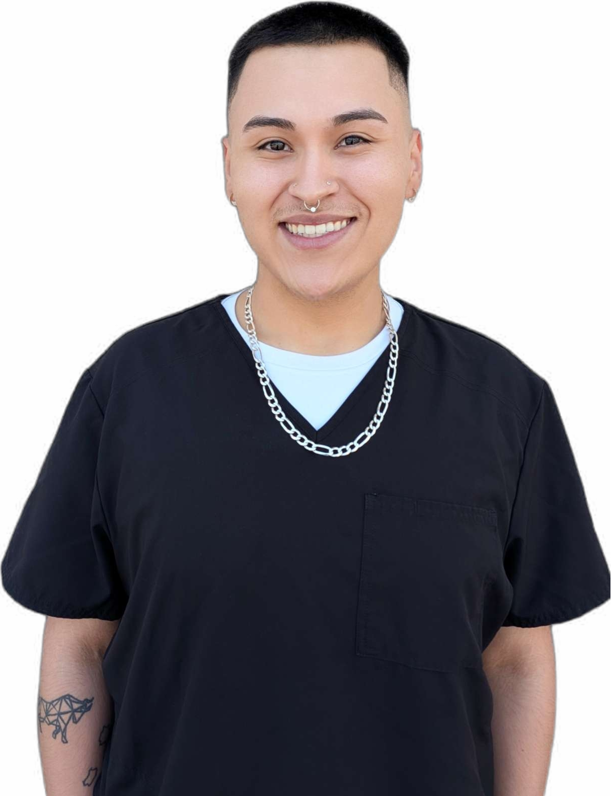 A person with short black hair, wearing a black medical scrub top, a white undershirt, a silver chain necklace, multiple earrings, and a septum piercing, smiling in front of a black background.