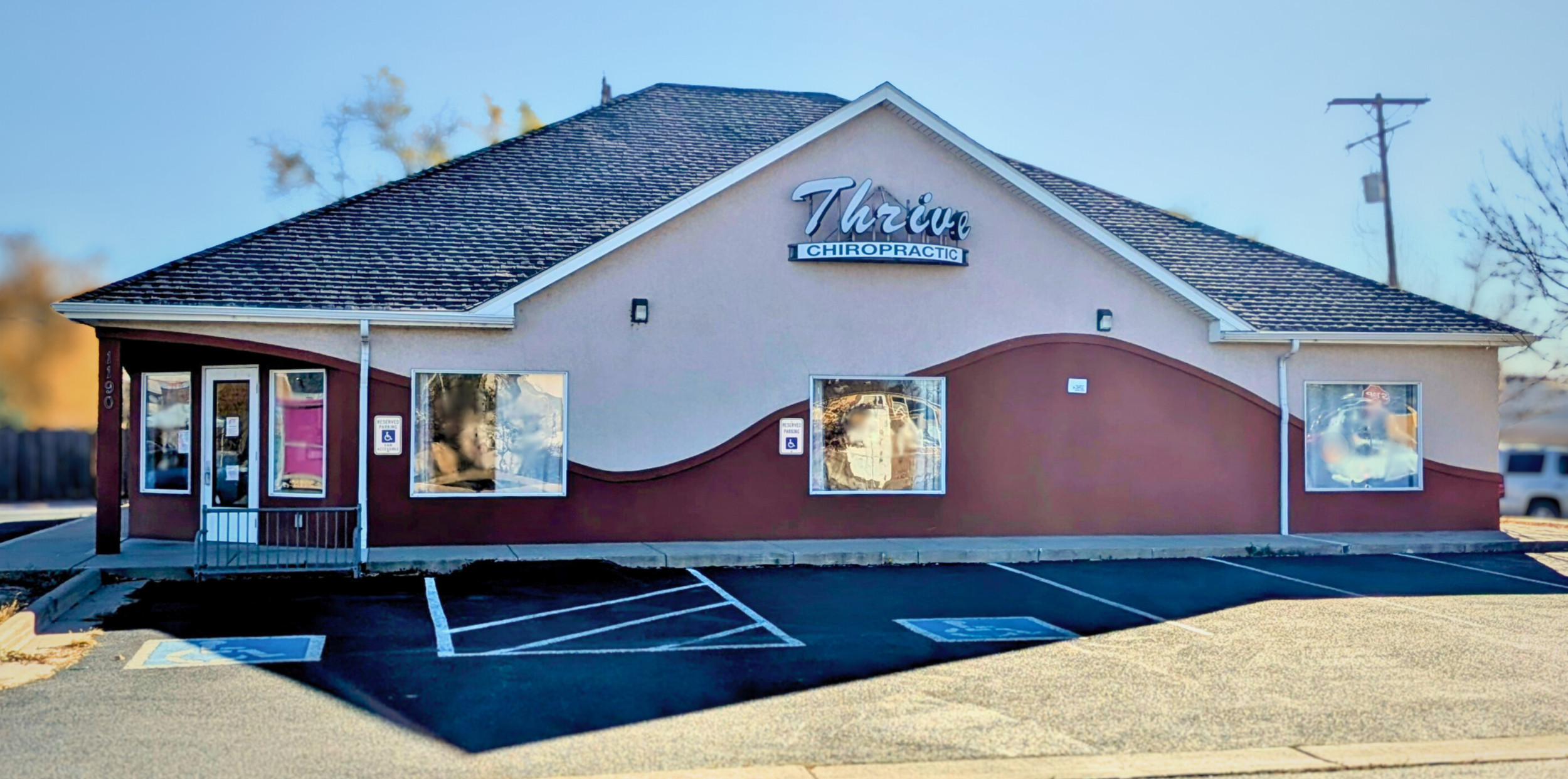 A small chiropractic clinic named 'Thrive Chiropractic' in a beige building with a dark red curved accent wall. The building has three large windows, a glass door, and reserved handicapped parking spots in front.