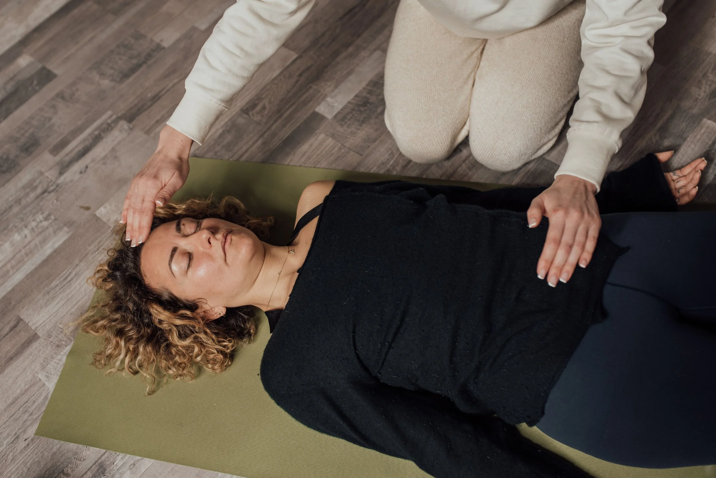 A woman with curly hair lying on a yoga mat with eyes closed, practicing relaxation or meditation, while another person kneels beside her with hands on her head.