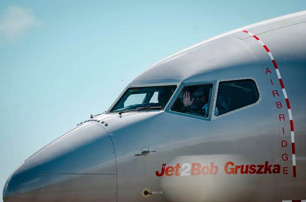 Close-up of a jet airplane's cockpit with a pilot waving, showing aircraft markings and the name 'Jet 2 Bob Gruszka' on the fuselage.