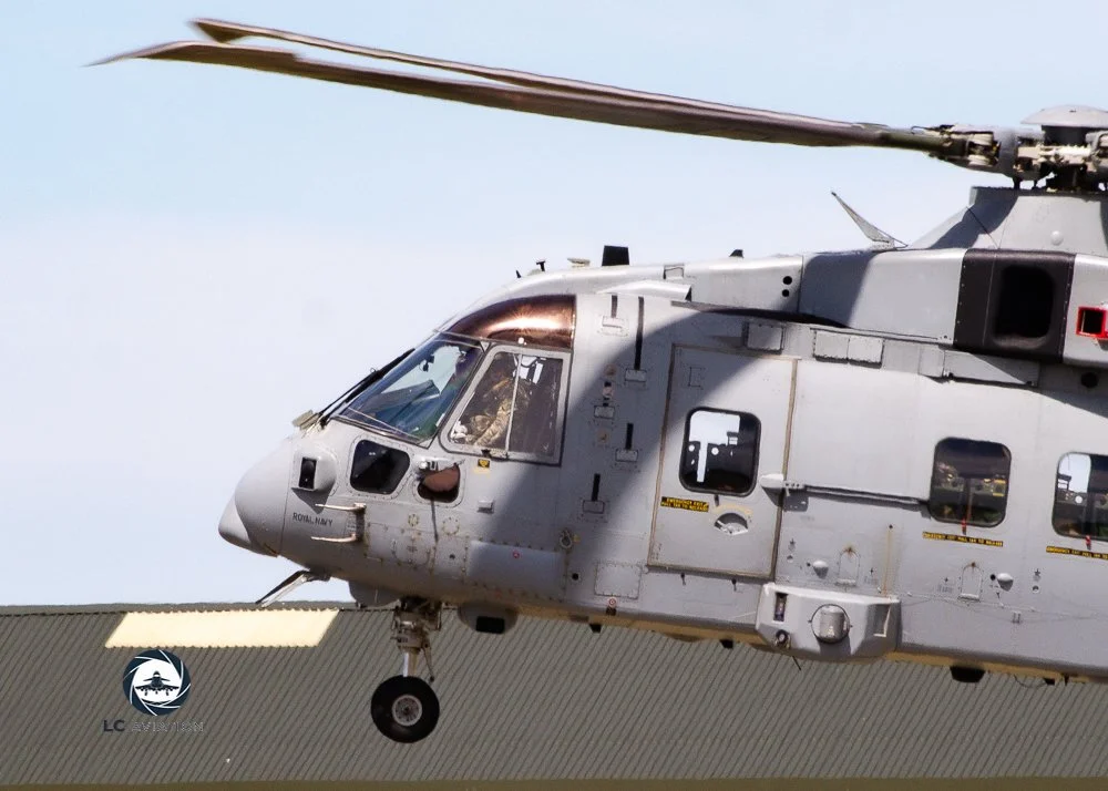 Close-up of a gray military helicopter in flight, with the pilot visible through the cockpit window, and the landing gear extended.