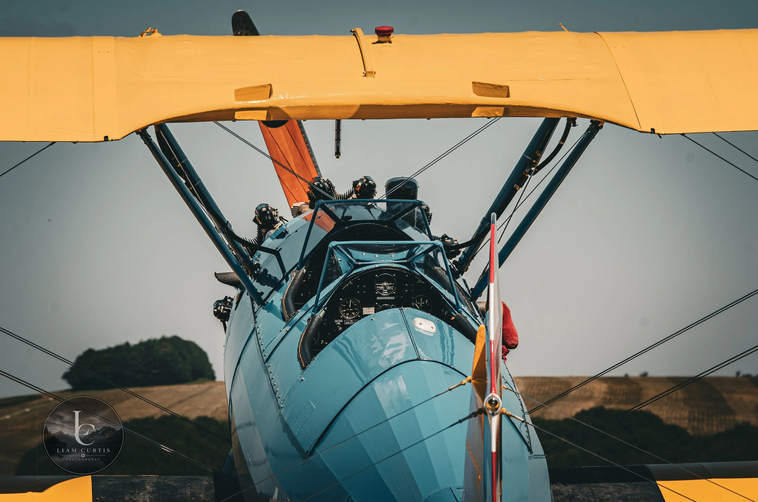 The image shows the front view of a vintage biplane aircraft with a light blue body and yellow upper wings, set against a background of a clear sky and distant hills.