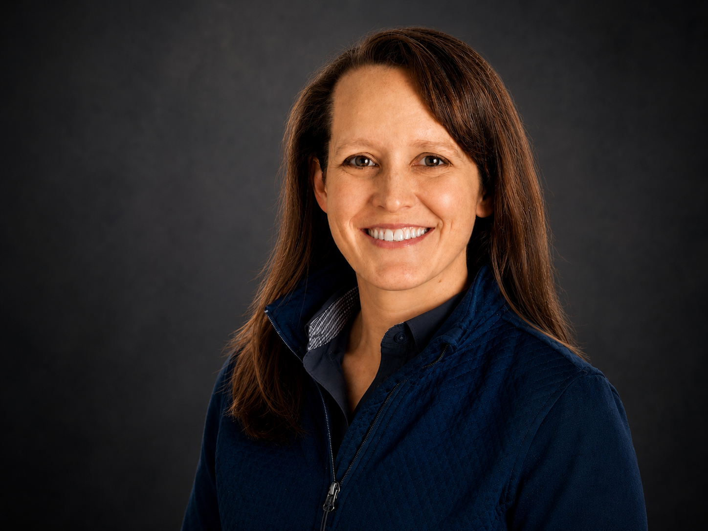 A woman with long brown hair smiling at the camera, wearing a dark blue jacket and a black shirt, against a dark background.