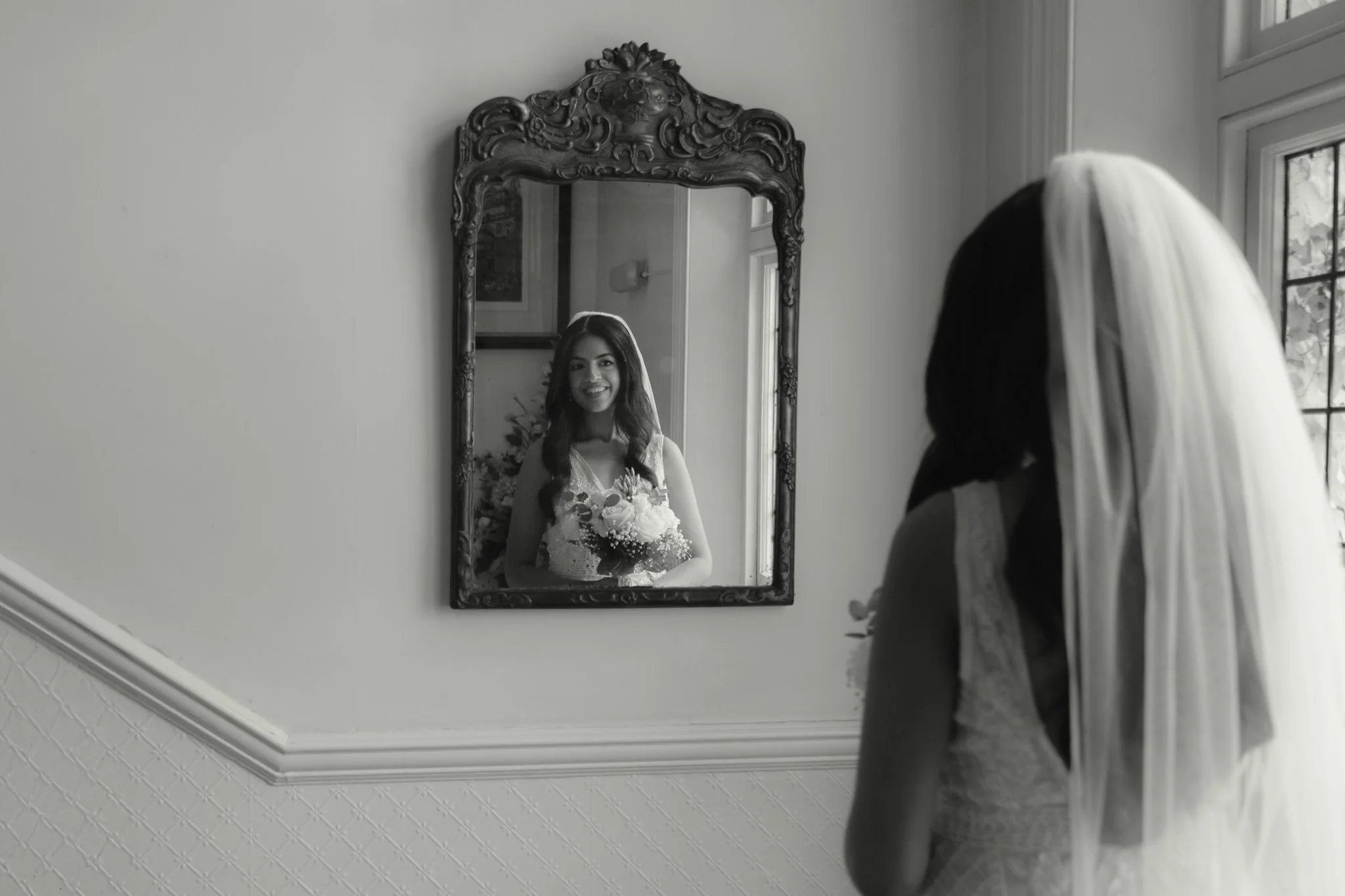 A young woman in wedding attire looking at herself in an ornate wall mirror, smiling, holding a bouquet of flowers, with a veil on her head, reflected in a black and white photo.