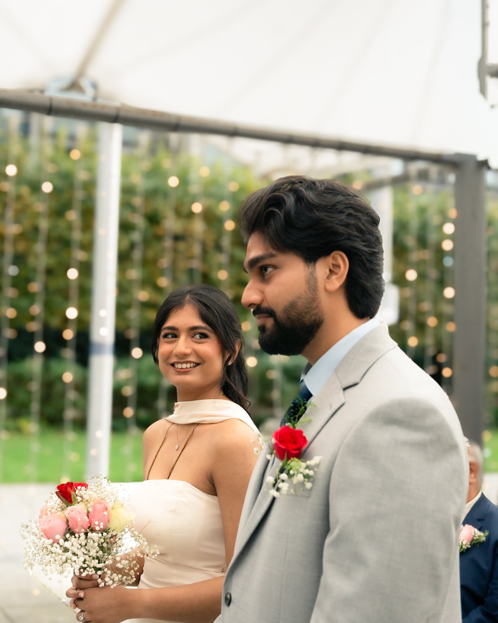 A bride and groom at a wedding ceremony outdoors, standing close, with the bride holding a bouquet of pink and red flowers, and the groom wearing a light gray suit with a red boutonniere, with a decorative backdrop of greenery and string lights.
