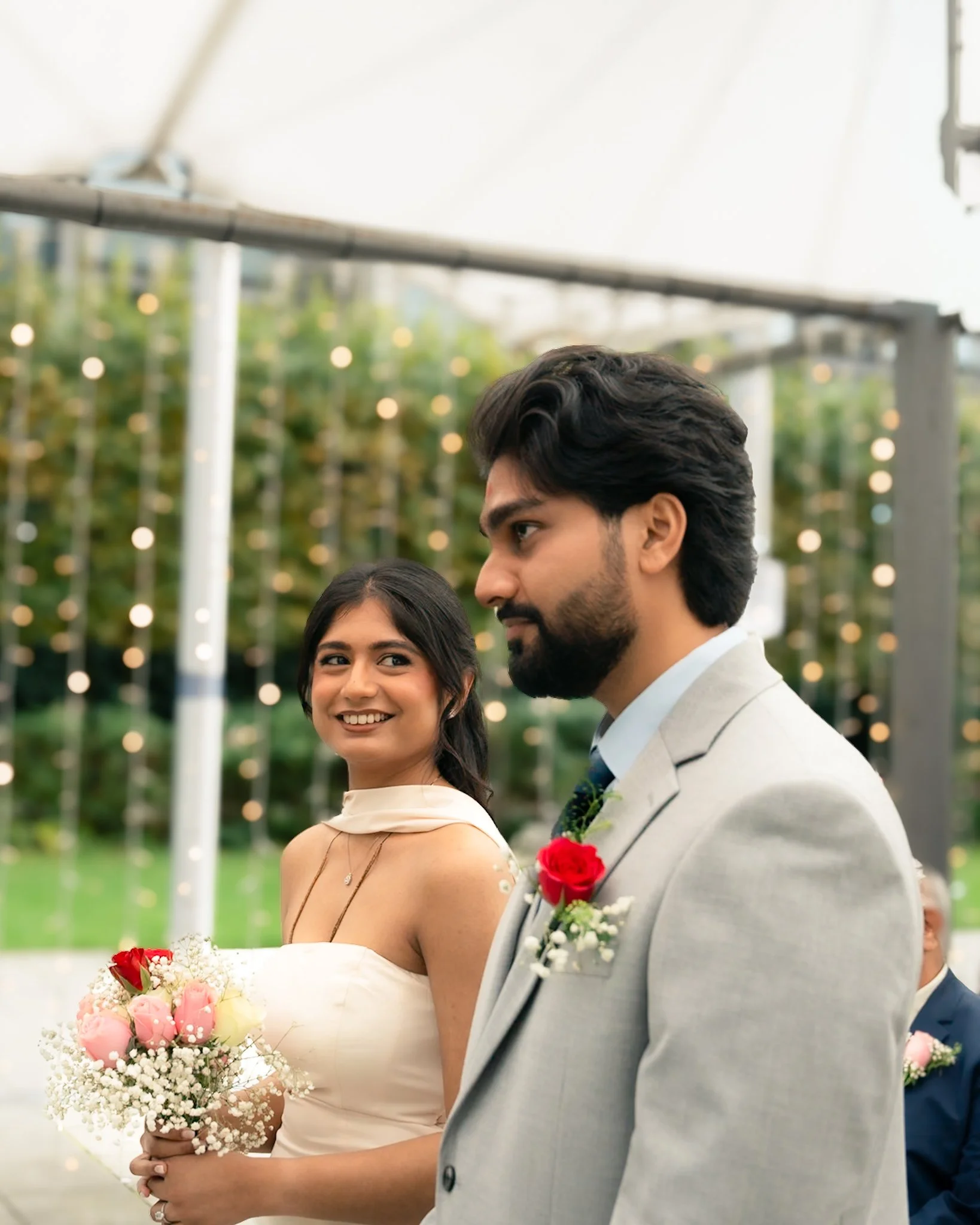 A happy bride and groom standing outdoors during their wedding ceremony, with the bride holding a bouquet of pink and white flowers, and the groom wearing a gray suit with a red rose boutonniere.