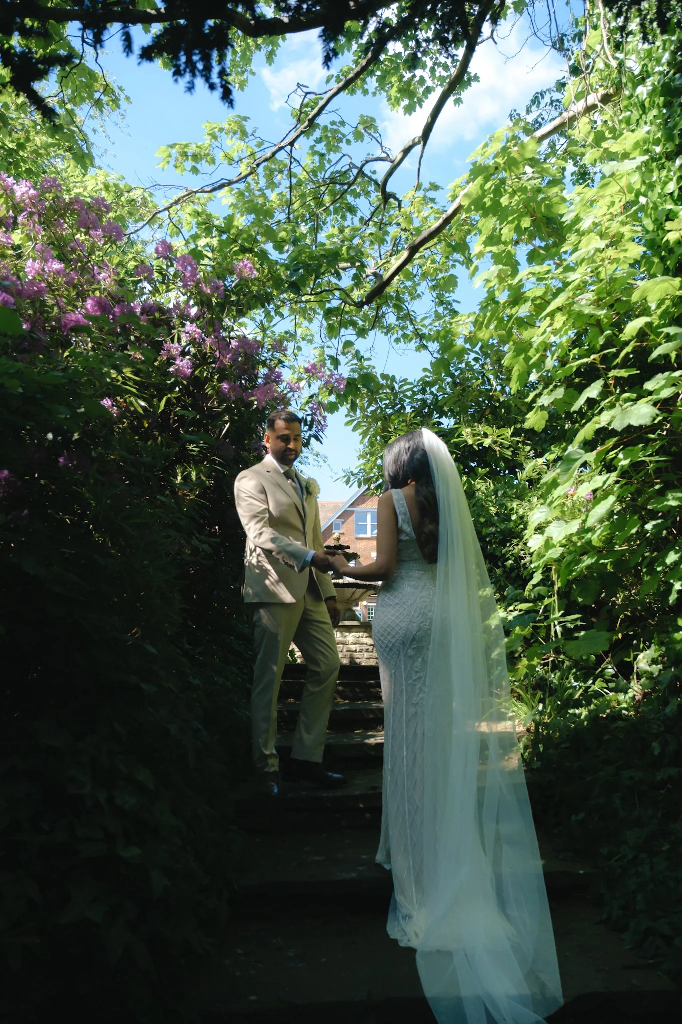 A wedding ceremony taking place outdoors on a staircase surrounded by greenery and trees, with a man and woman exchanging vows. The man is dressed in a light beige suit, and the woman is wearing a white wedding dress with a long veil.