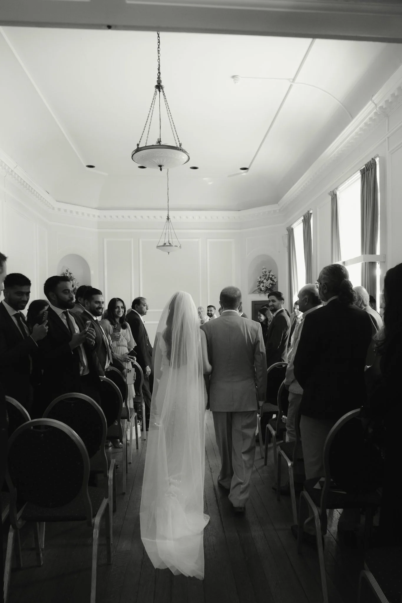 A black and white photo of a wedding ceremony in a large, ornate room with high ceilings and large windows. The bride, dressed in a long white gown and veil, faces the groom, dressed in a light-colored suit. They are surrounded by guests standing and