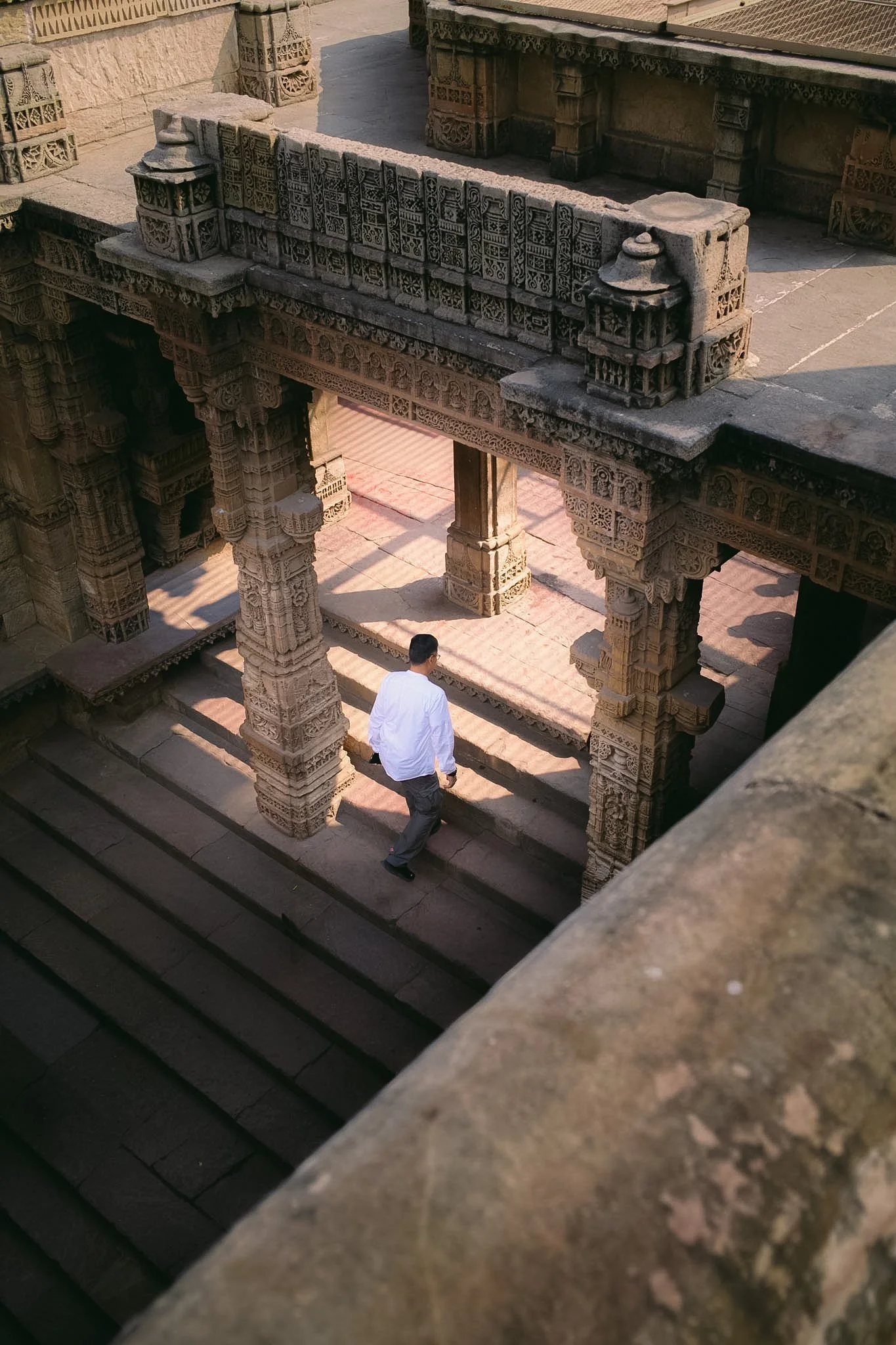 The image shows a man in a white shirt walking down stone steps of a historic temple with intricate carvings, possibly in India, viewed from above.