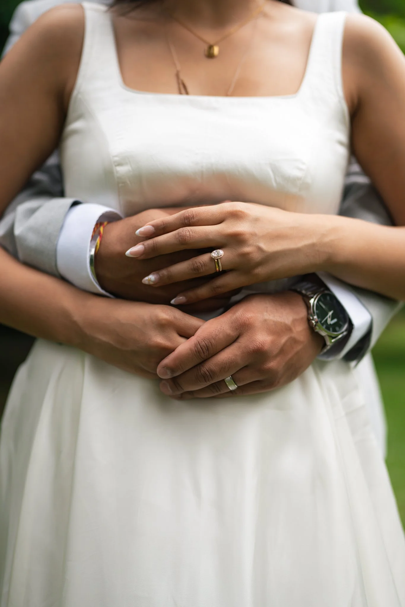 Close-up of a woman's torso, showing her wearing a white dress, jewelry, and a ring, with someone else's hands placed over her waist.